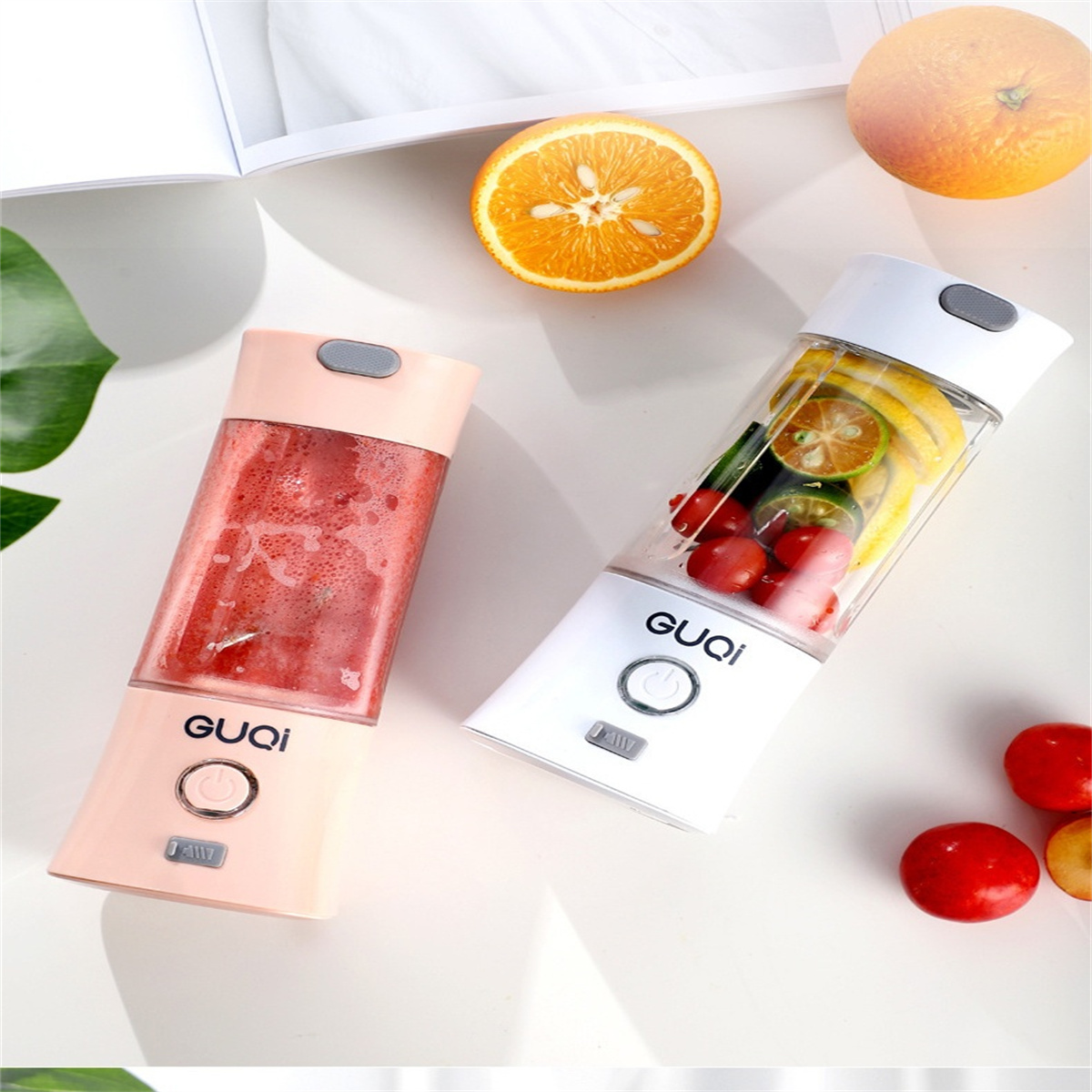 Zwei GUQi-Mixer, einer rosa mit Erdbeeren, der andere weiß mit Orangen und Limetten, auf weißer Oberfläche.