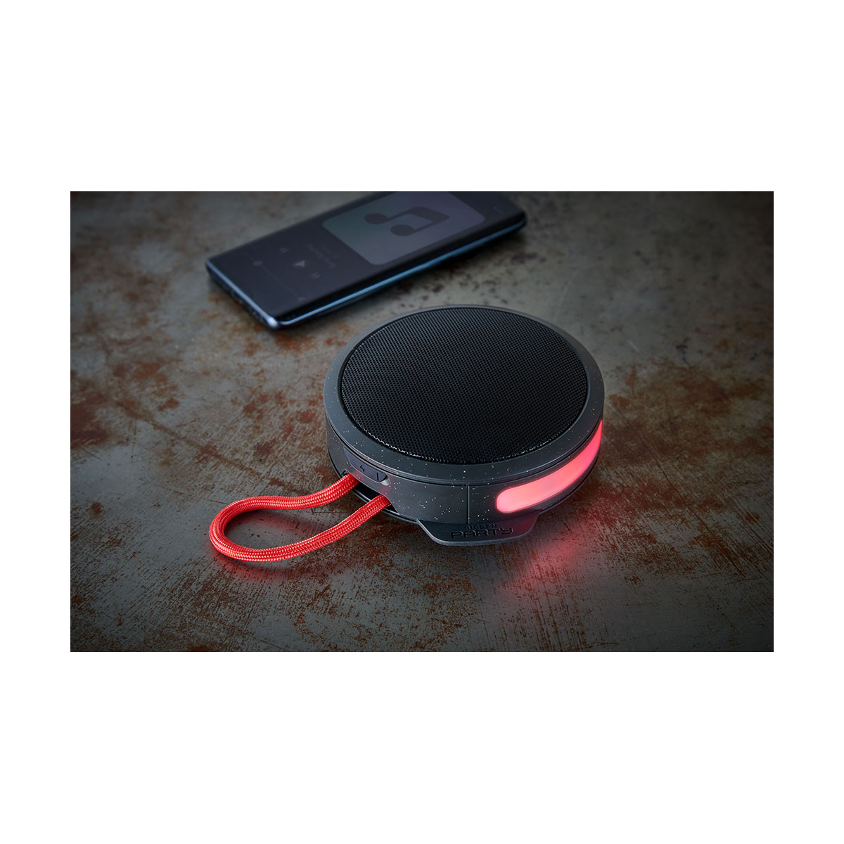 Czarny okrągły głośnik z czerwonym paskiem i czerwonymi światłami. Głośnik znajduje się na powierzchni.