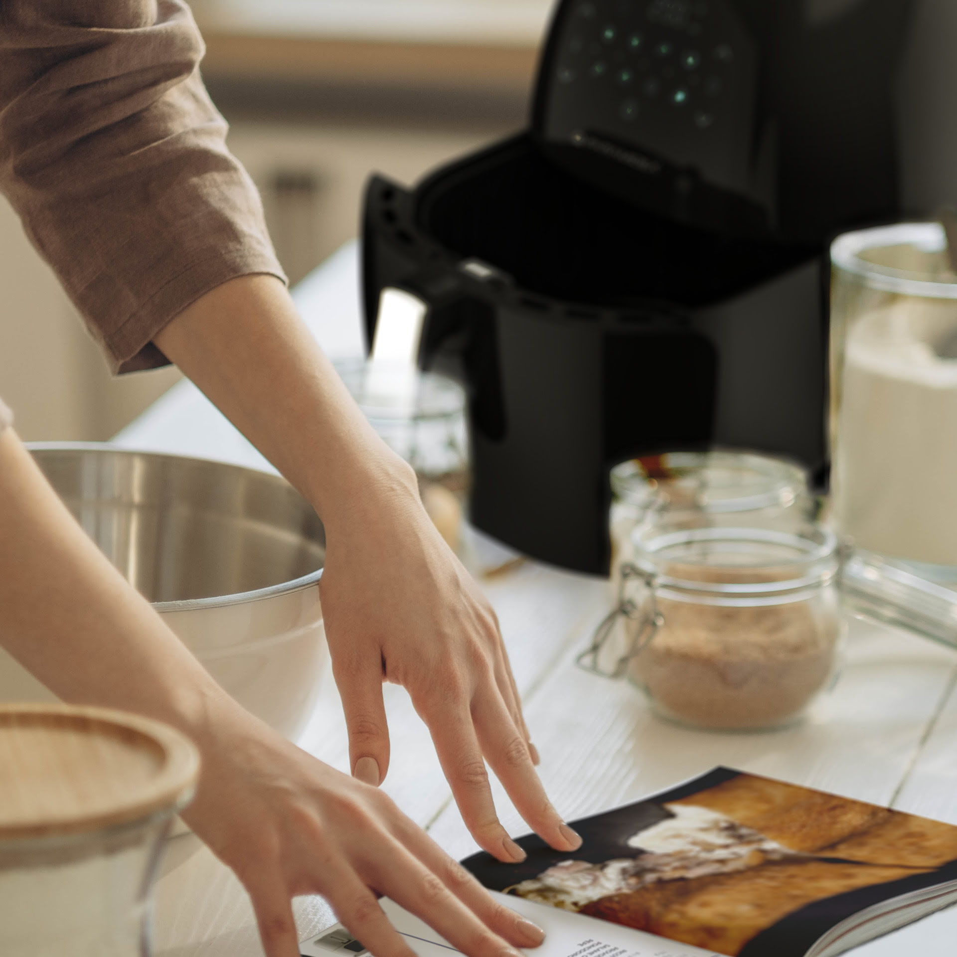 Le mani di una persona sono vicino a un libro di cucina aperto e a una friggitrice ad aria nera.