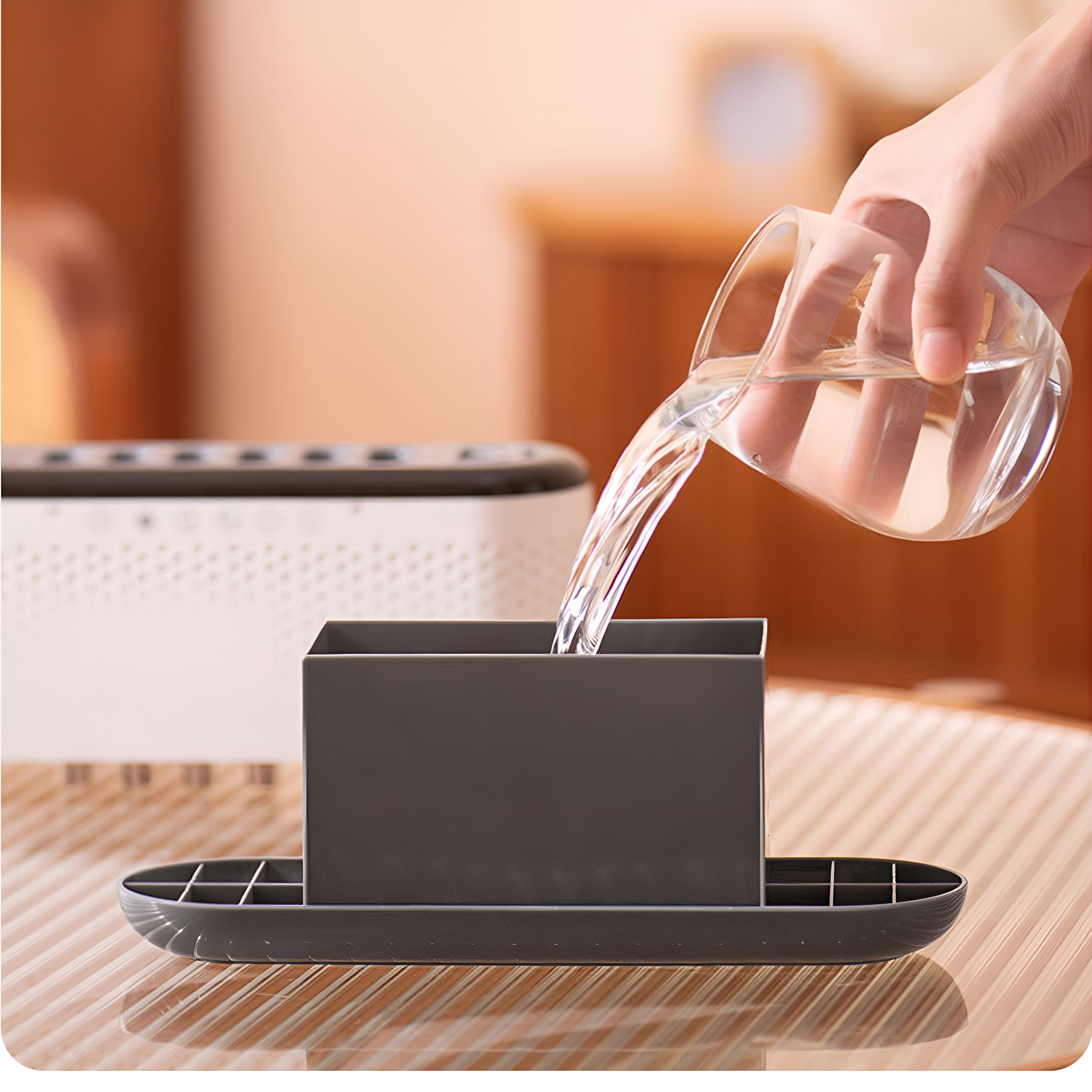 Eine Hand gießt Wasser in einen dunkelgrauen Behälter, der auf einem Tablett steht. Ein weißer Luftreiniger im Hintergrund.