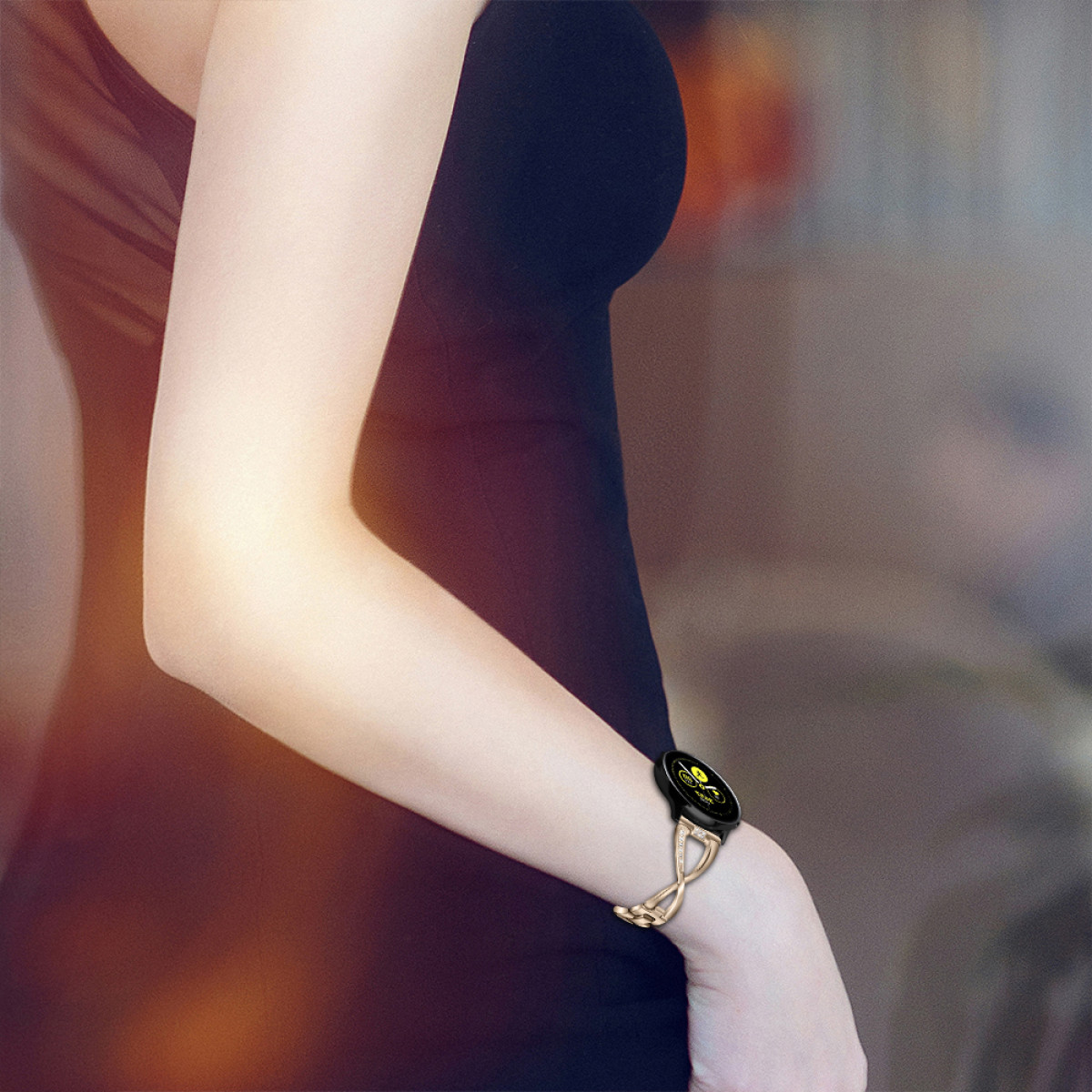 Ramię kobiety ze złotym zegarkiem, w czarnej sukience, rozmyte tło.