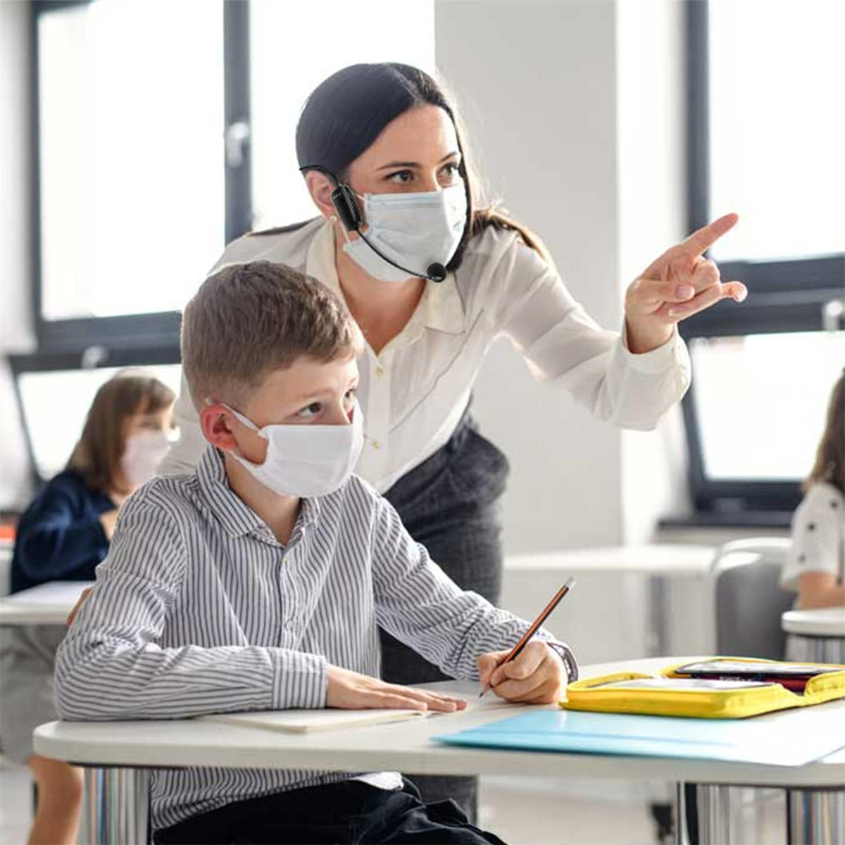 Eine Lehrerin mit Maske und Headset zeigt auf einen Schüler mit Maske im Klassenzimmer.