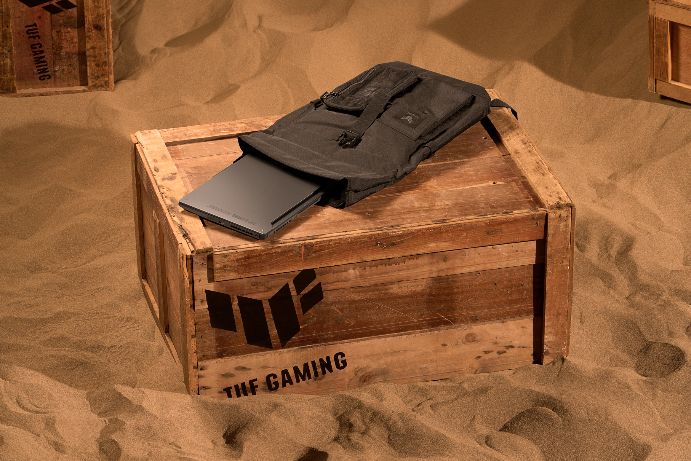 Laptop in einem schwarzen Rucksack auf einer Holzkiste. Die Kiste hat ein Design. Die Szene spielt in einer sandigen Umgebung.