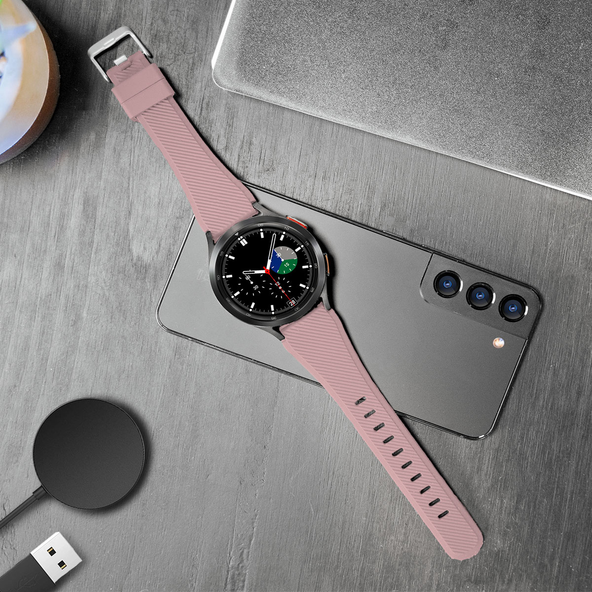 Smartwatch auf einem Tisch neben Telefon und Ladepad. Mit pinkfarbenem Armband.