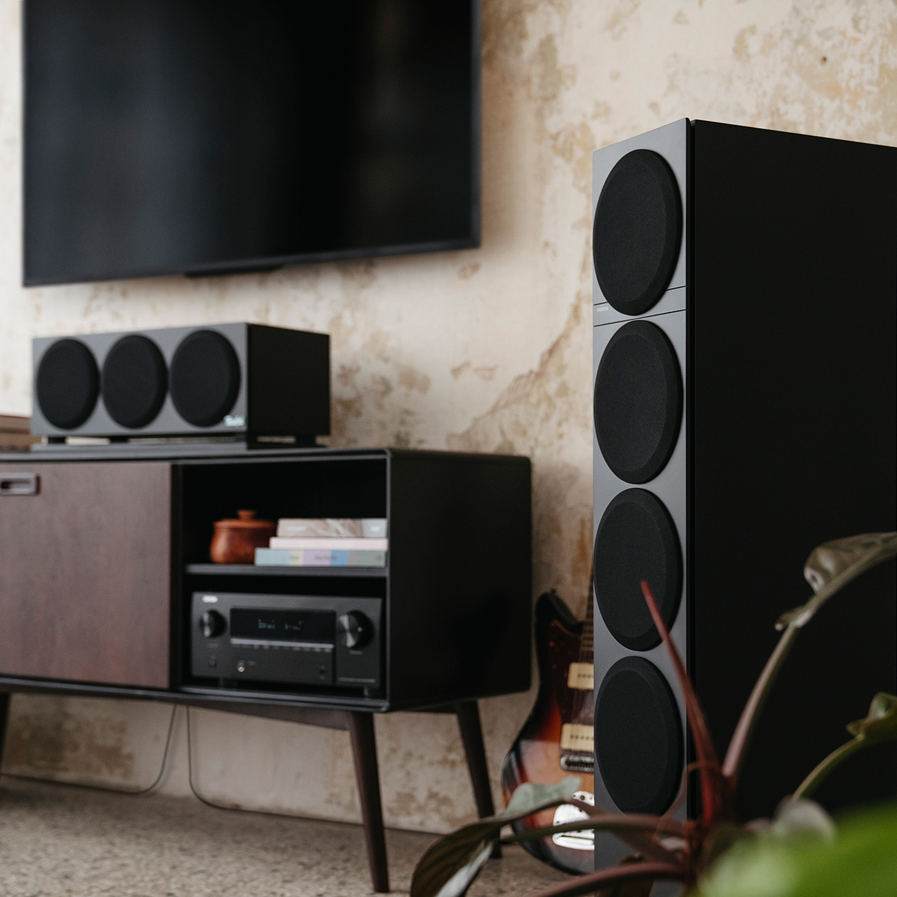 Wohnzimmer-Setup mit Fernseher, Lautsprechern und einem Holzschrank. Eine Gitarre lehnt an einem Lautsprecher.