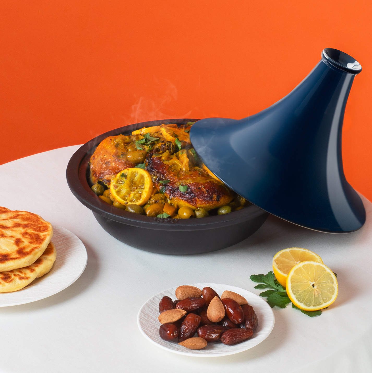Un tagine marocchino con cibo fumante, circondato da contorni e limoni, su una tovaglia bianca.