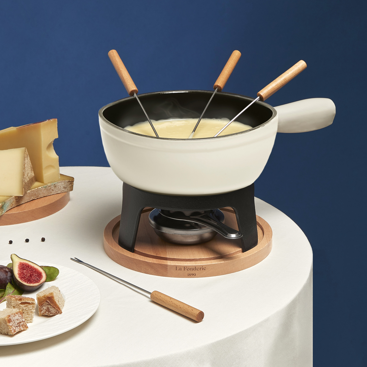 Set per fonduta con formaggio, pane e fichi su un tavolo bianco e sfondo blu.