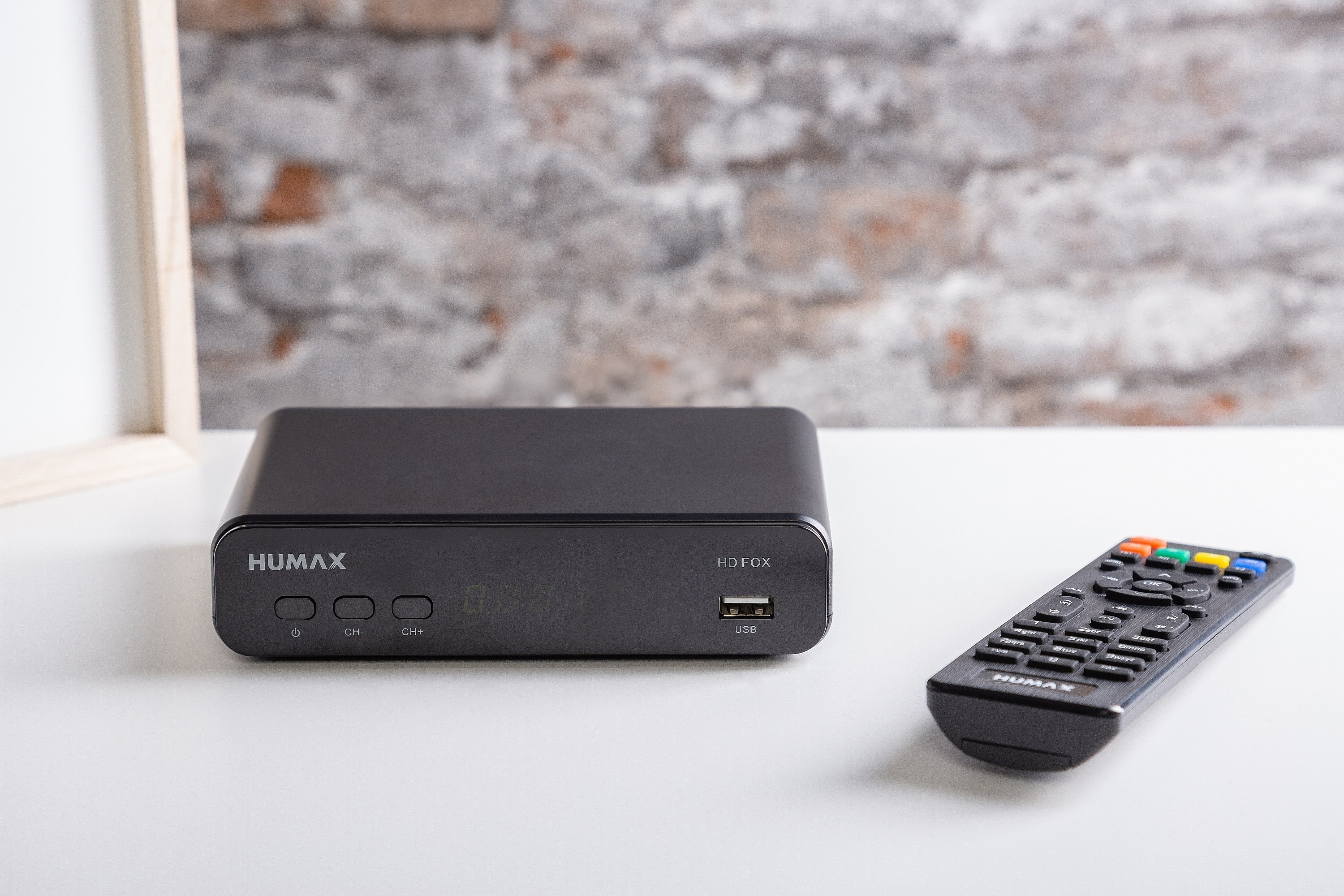 Schwarze Set-Top-Box mit Fernbedienung. Die Box hat einen USB-Anschluss und ein Display. Weißer Tisch.