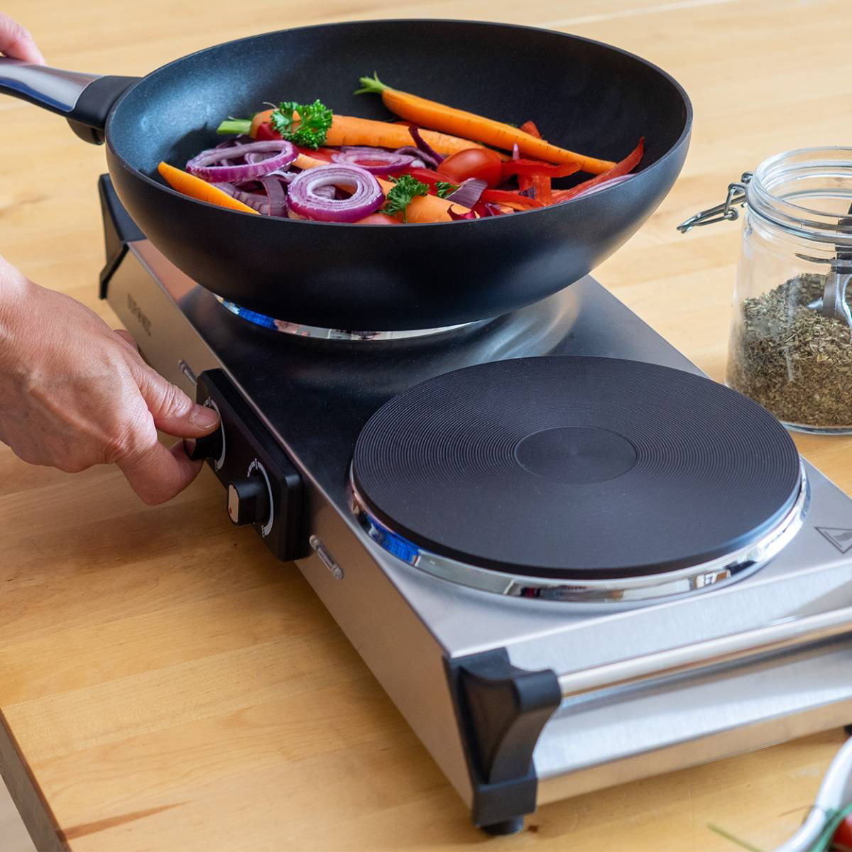 Een persoon kookt groenten in een pan. De pan staat op een kookplaat op een houten oppervlak.
