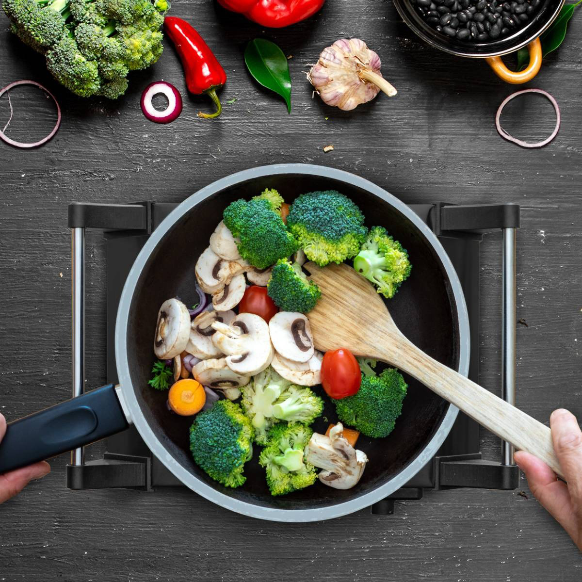 Une poêle avec légumes et une spatule en bois sur une plaque, ingrédients autour.