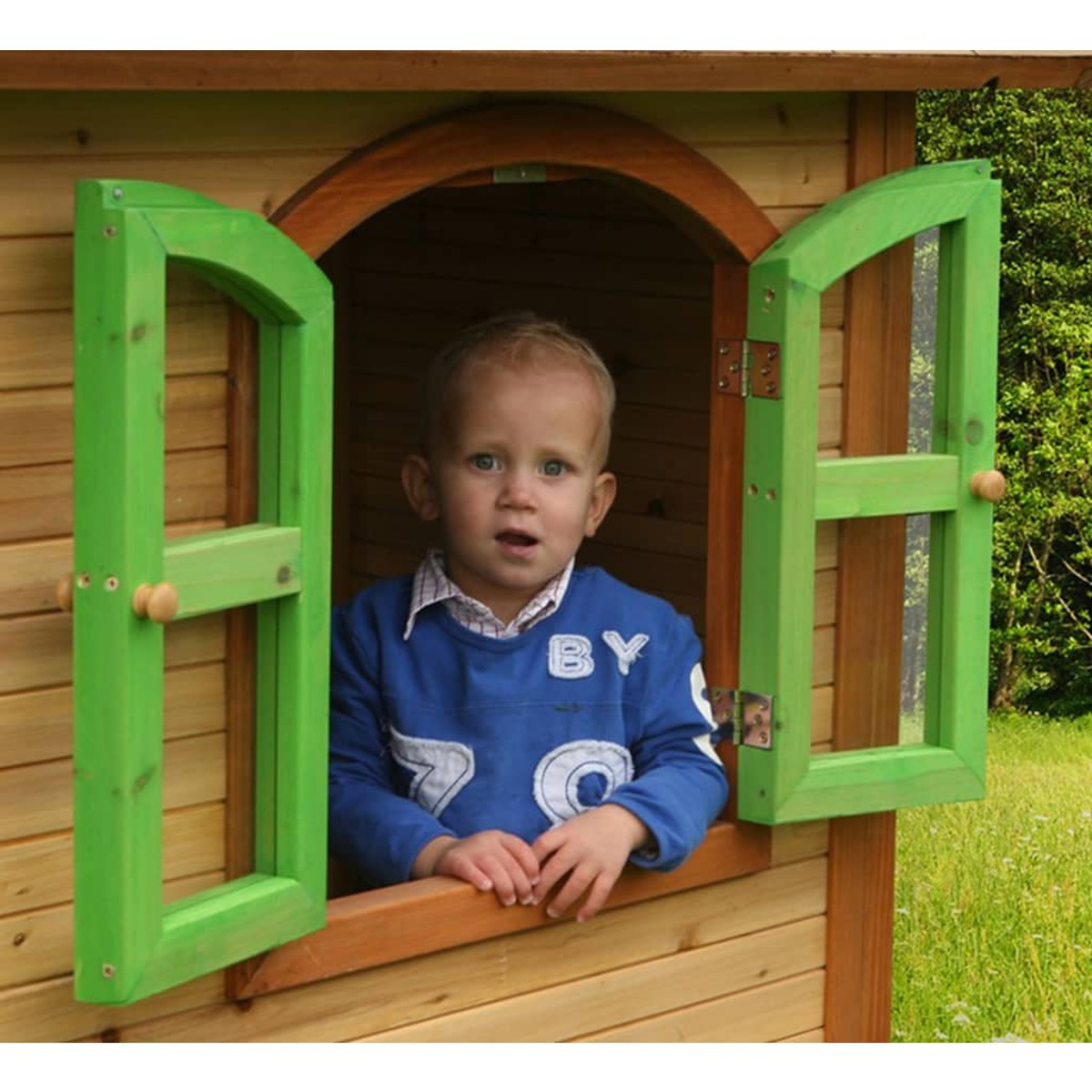 Ein kleiner Junge schaut aus einem Holzspielhausfenster. Der Fensterrahmen ist grün.