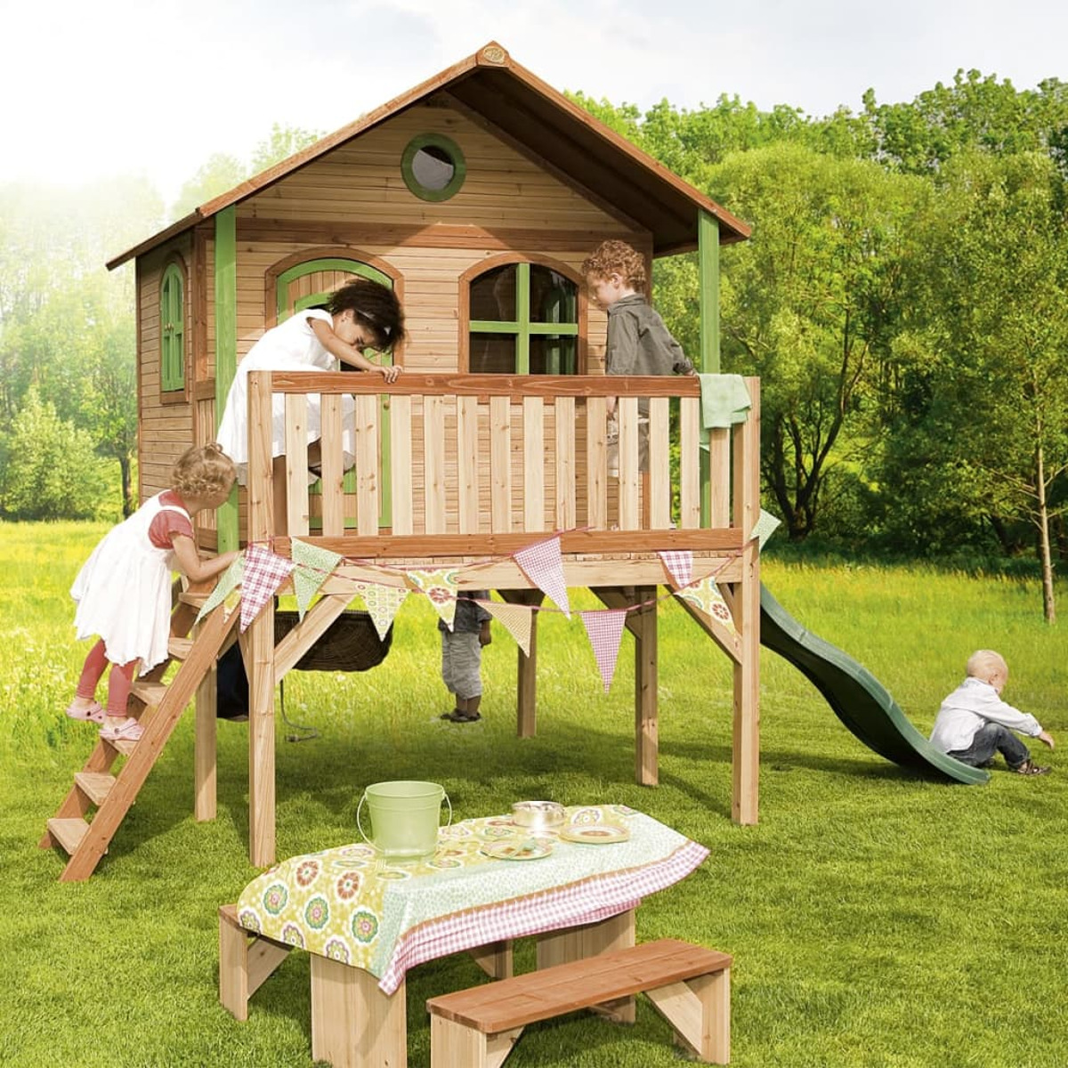 Kinder spielen in einem Holzspielhaus mit Rutsche. Grünes Gras und blauer Himmel.