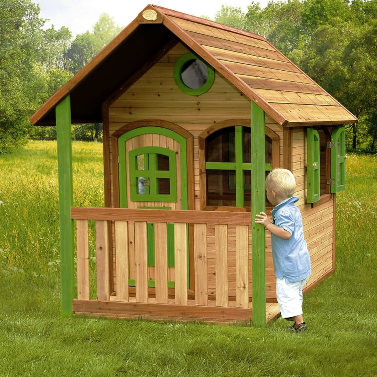 Ein Holzspielhaus mit grünen Akzenten. Ein Junge in Blau betrachtet das Haus in einer grasbewachsenen Gegend mit Bäumen.
