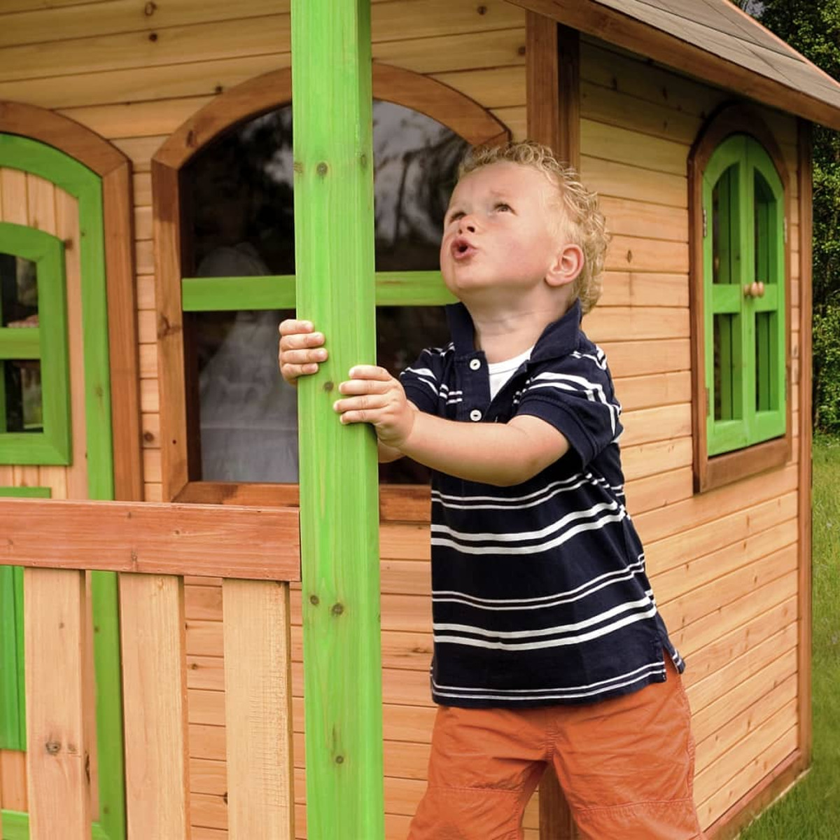 Ein kleiner Junge, der nach oben auf ein Spielhaus blickt und einen grünen Träger hält.