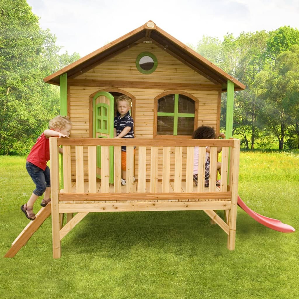Kinder spielen in einem Holzspielhaus mit Rutsche. Zwei Kinder sind auf der Terrasse und eines klettert.