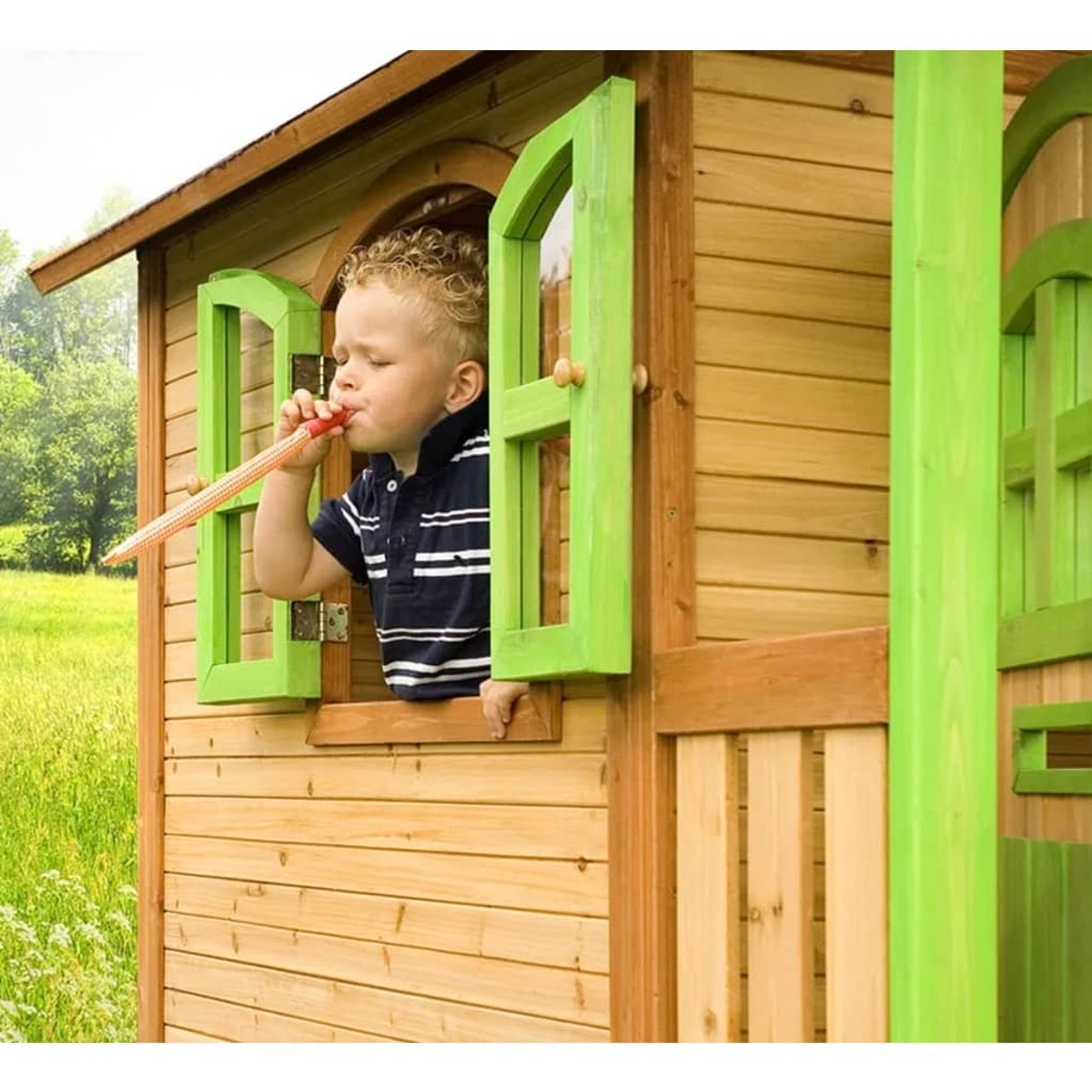 Junge in einem Spielhaus aus Holz, der durch ein Fenster in eine Partytröte bläst.