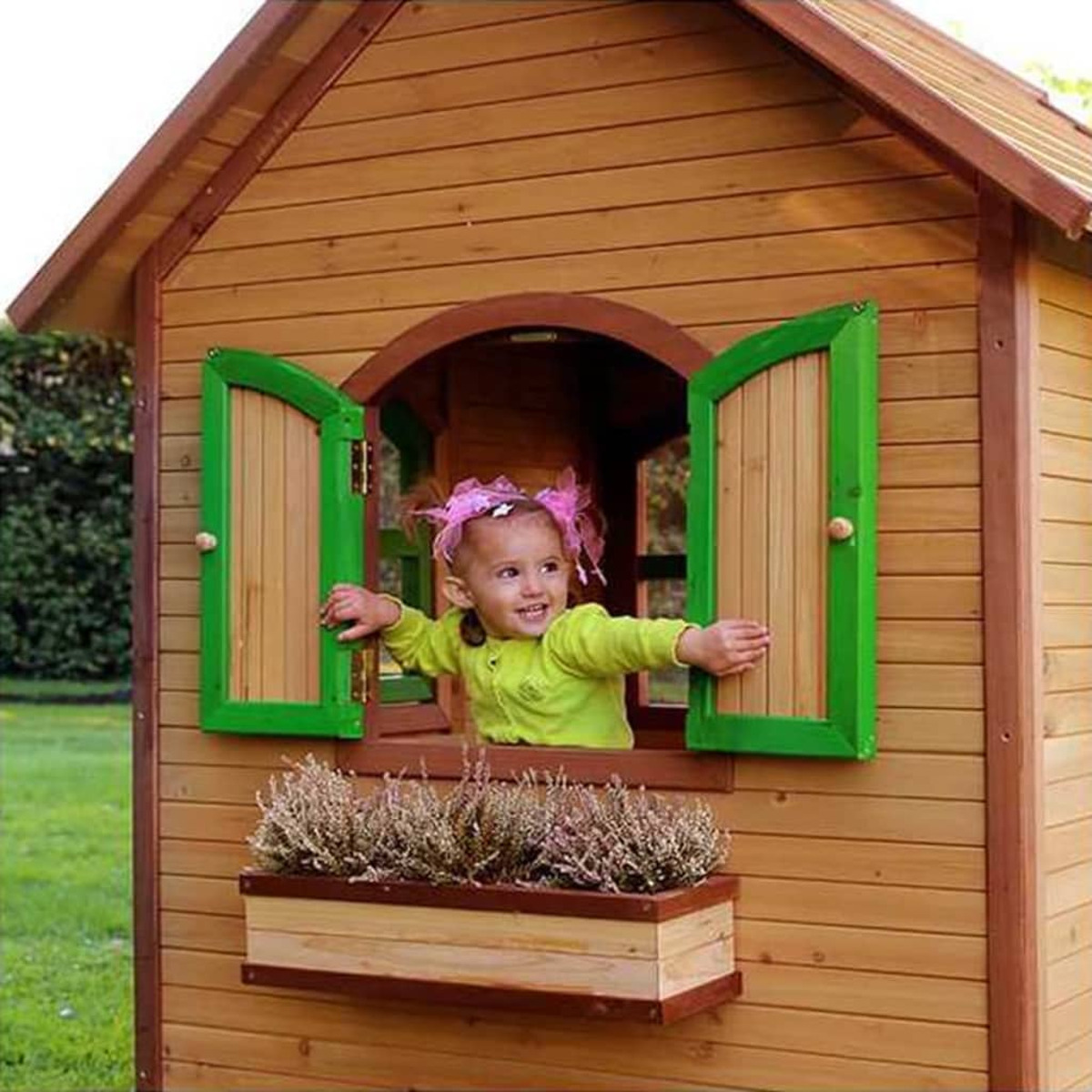 Ein Kind in einem hölzernen Spielhaus. Grüne Fensterläden und Blumenkasten sind sichtbar.