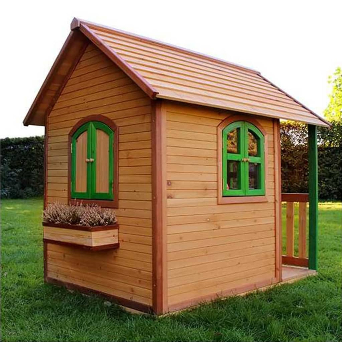 Ein Spielhaus aus Holz mit grünen Fenstern und Verzierungen. Das Haus steht auf Gras und zeigt eine Veranda und einen Blumenkasten.
