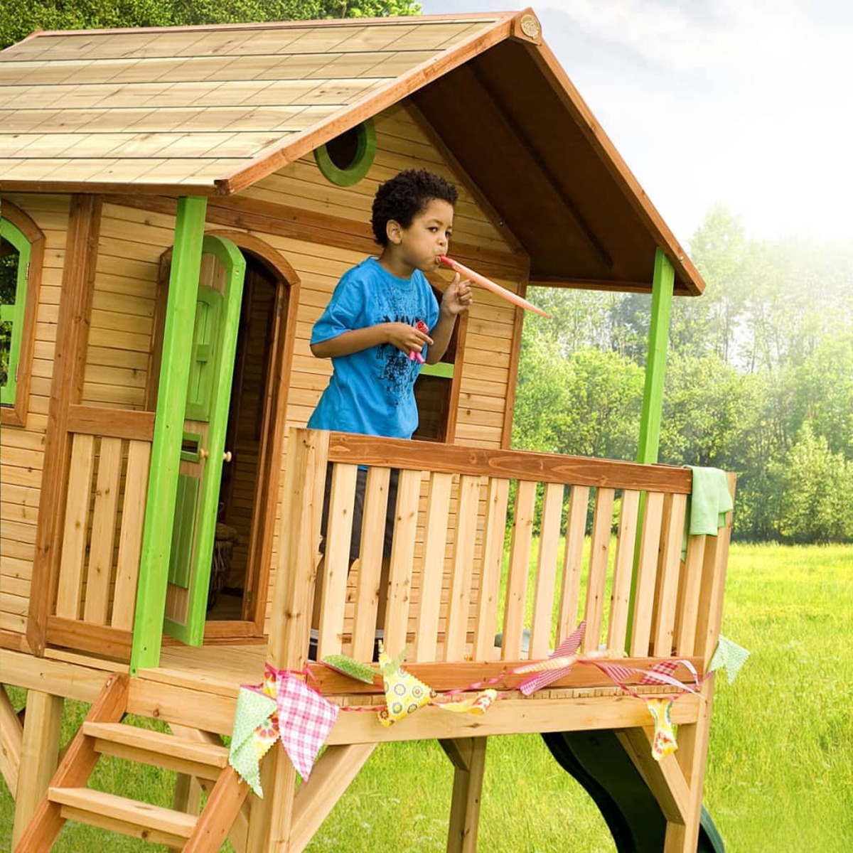 Kind auf Holzspielhaus-Balkon, das eine Partyhupe bläst, grünes Gras.