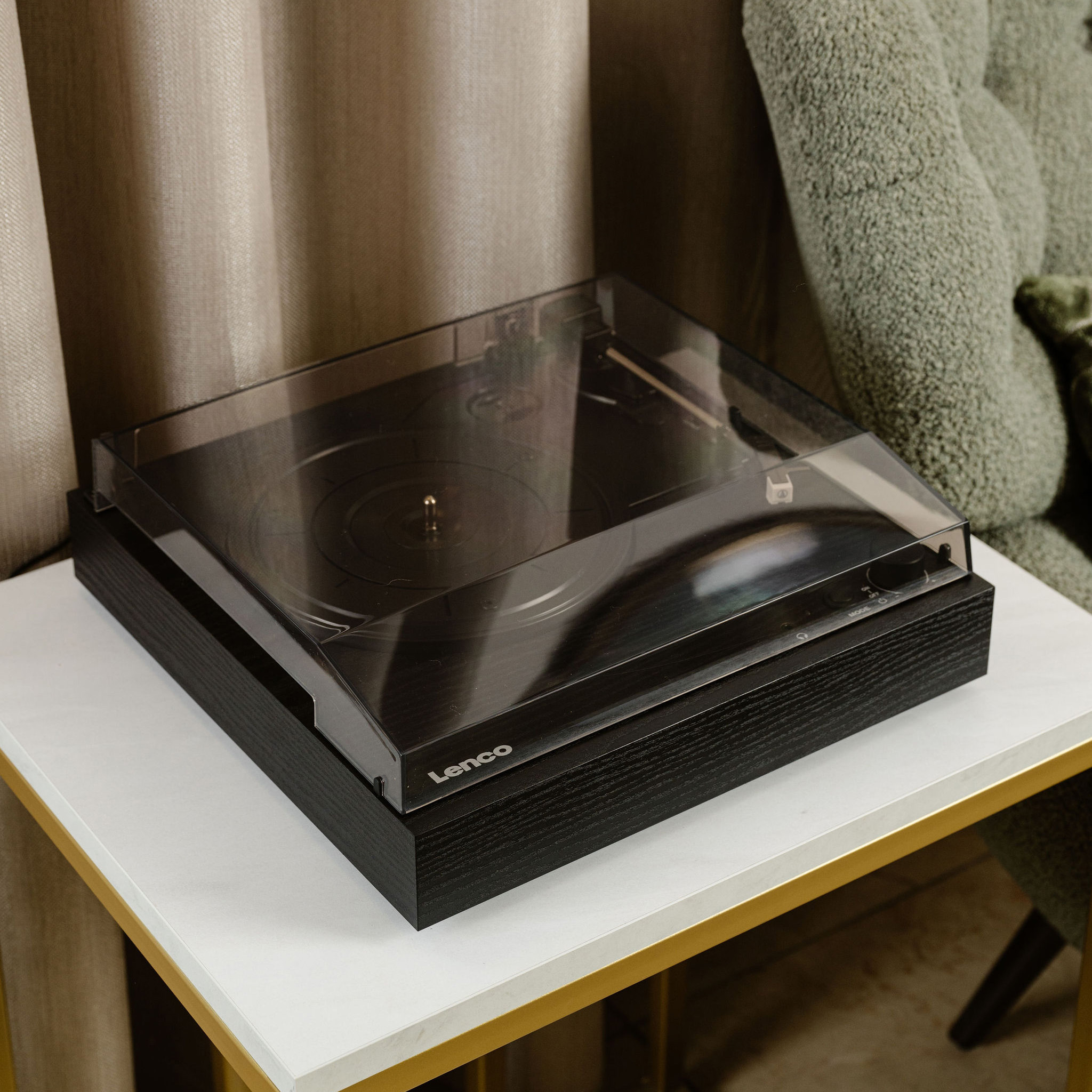 A black record player on a white table. The player has a clear lid and a wooden base, with a gold frame.