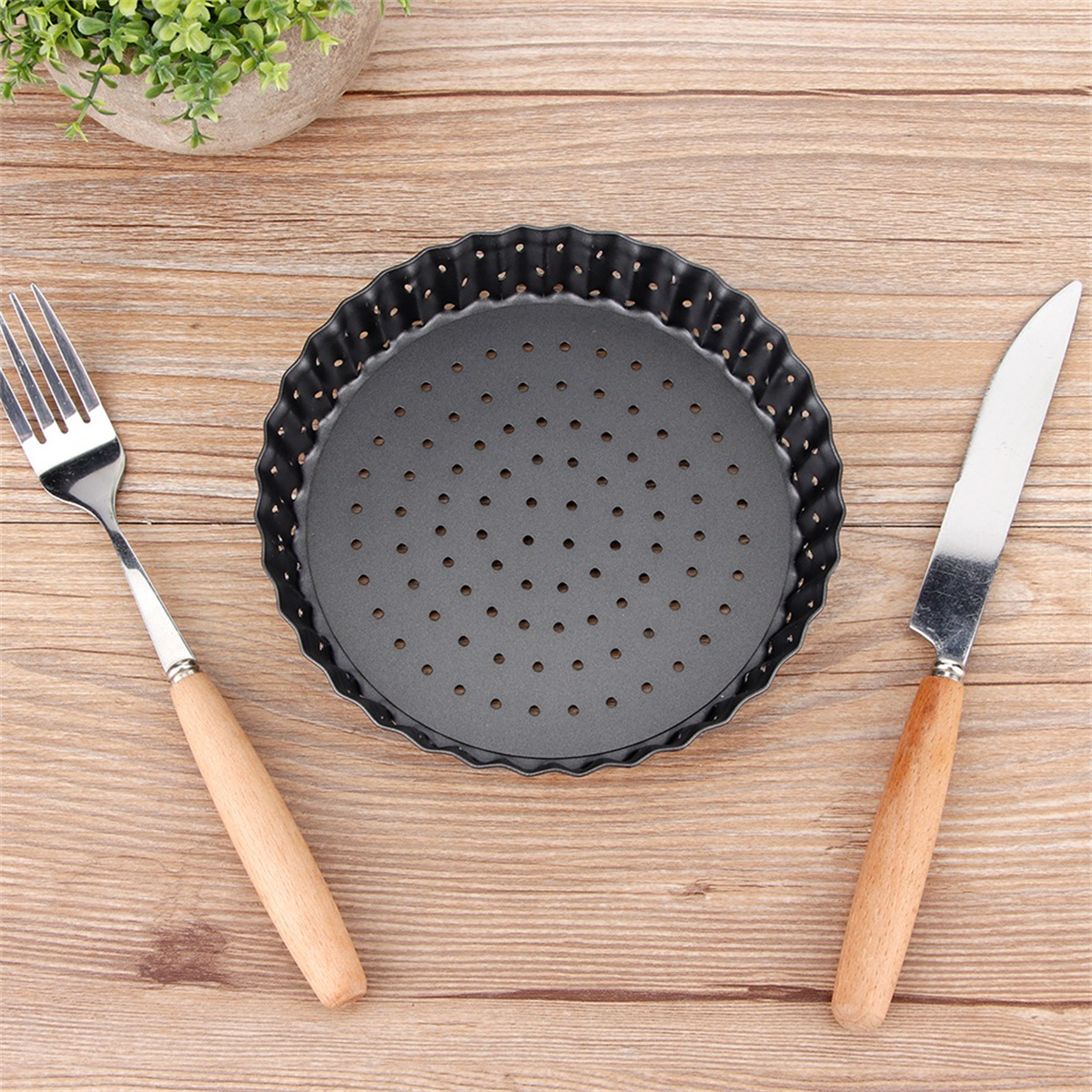 Runde schwarze Tarteform mit Löchern, eine Gabel und ein Messer mit Holzgriff. Auf Holz.