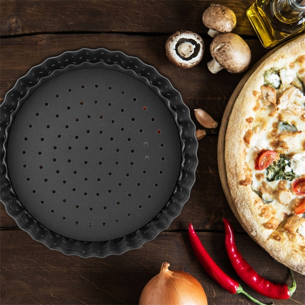 Eine runde, schwarze Pizzaform mit Löchern liegt auf einer Holzoberfläche mit Pilzen, Knoblauch, Chilischoten und einer Pizza.