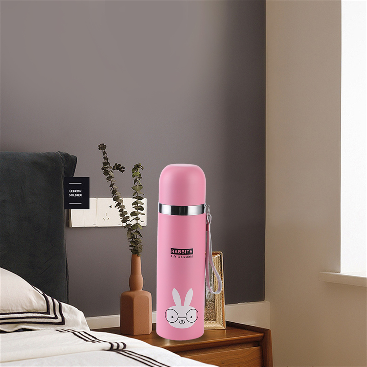 Rosa Thermoskanne mit Hasenmuster, auf einem Bett mit Vase und gerahmtem Bild platziert.