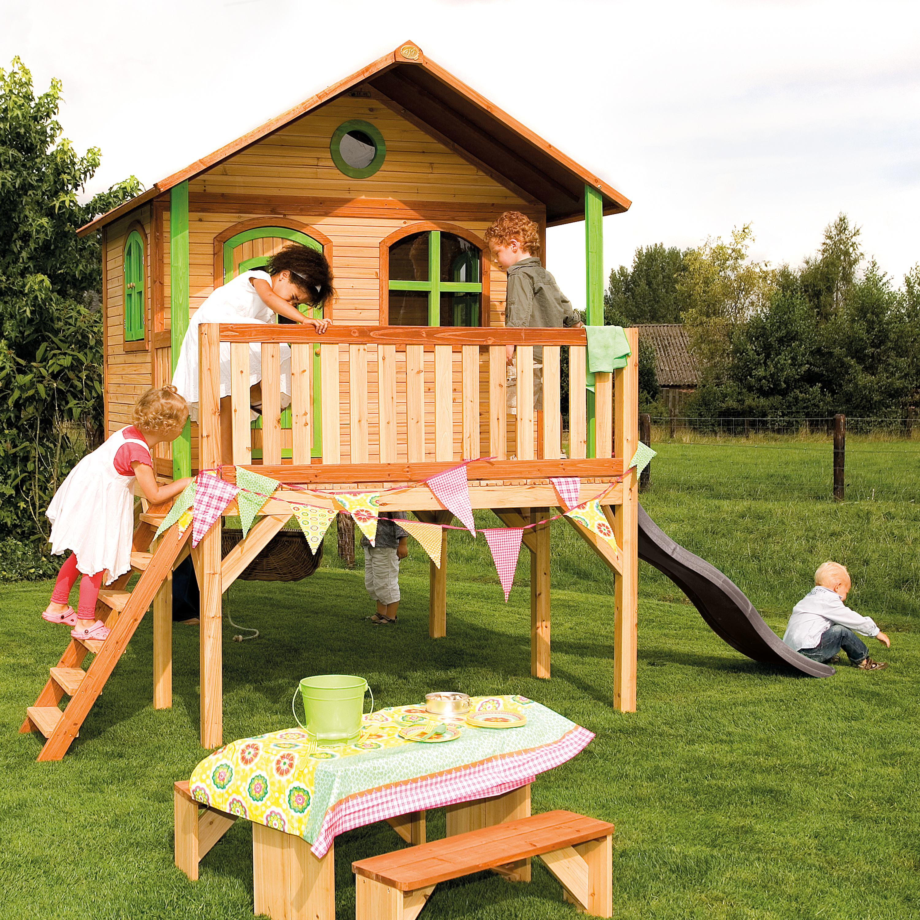 Kinder spielen auf einem Holzspielhaus mit Rutsche. Ein Tisch und eine Bank sind aufgebaut.