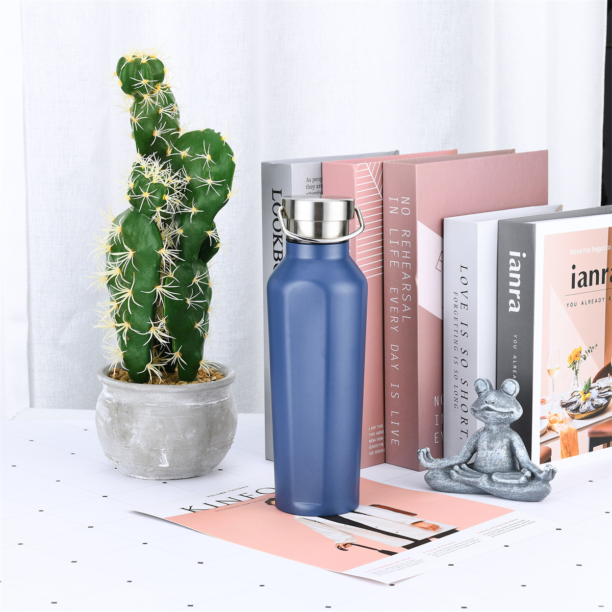 Eine blaue Wasserflasche neben einem Kaktus und Büchern. Eine kleine Froschstatue ist auch vorhanden.