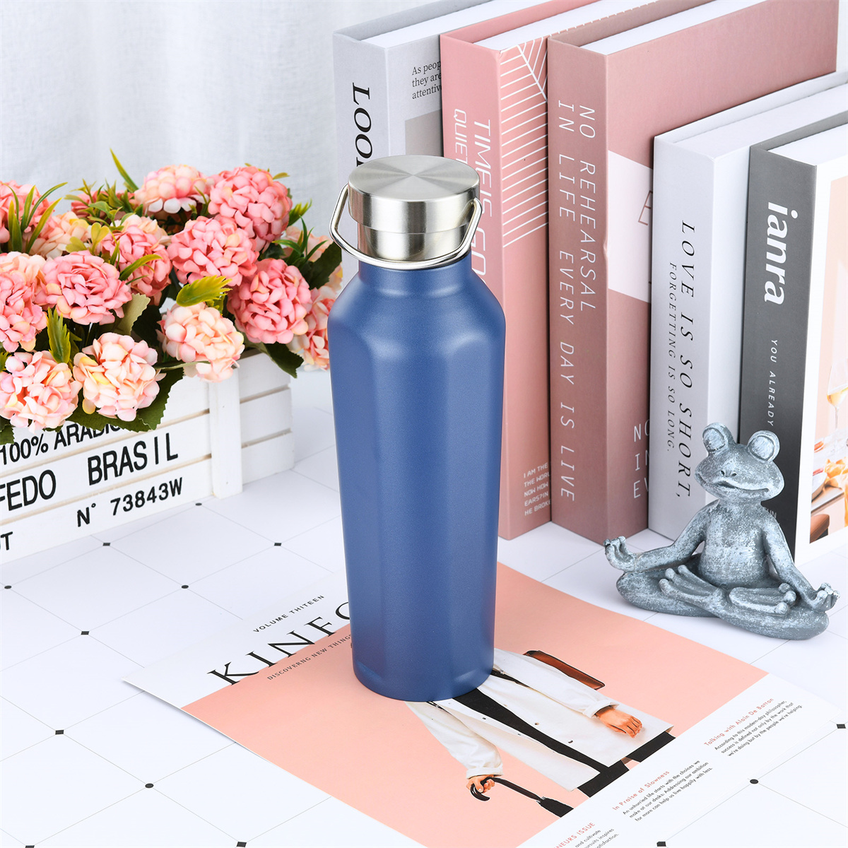 Blaue Wasserflasche mit silbernem Deckel auf einem Tisch. Es gibt Bücher, Blumen und eine kleine Statue.