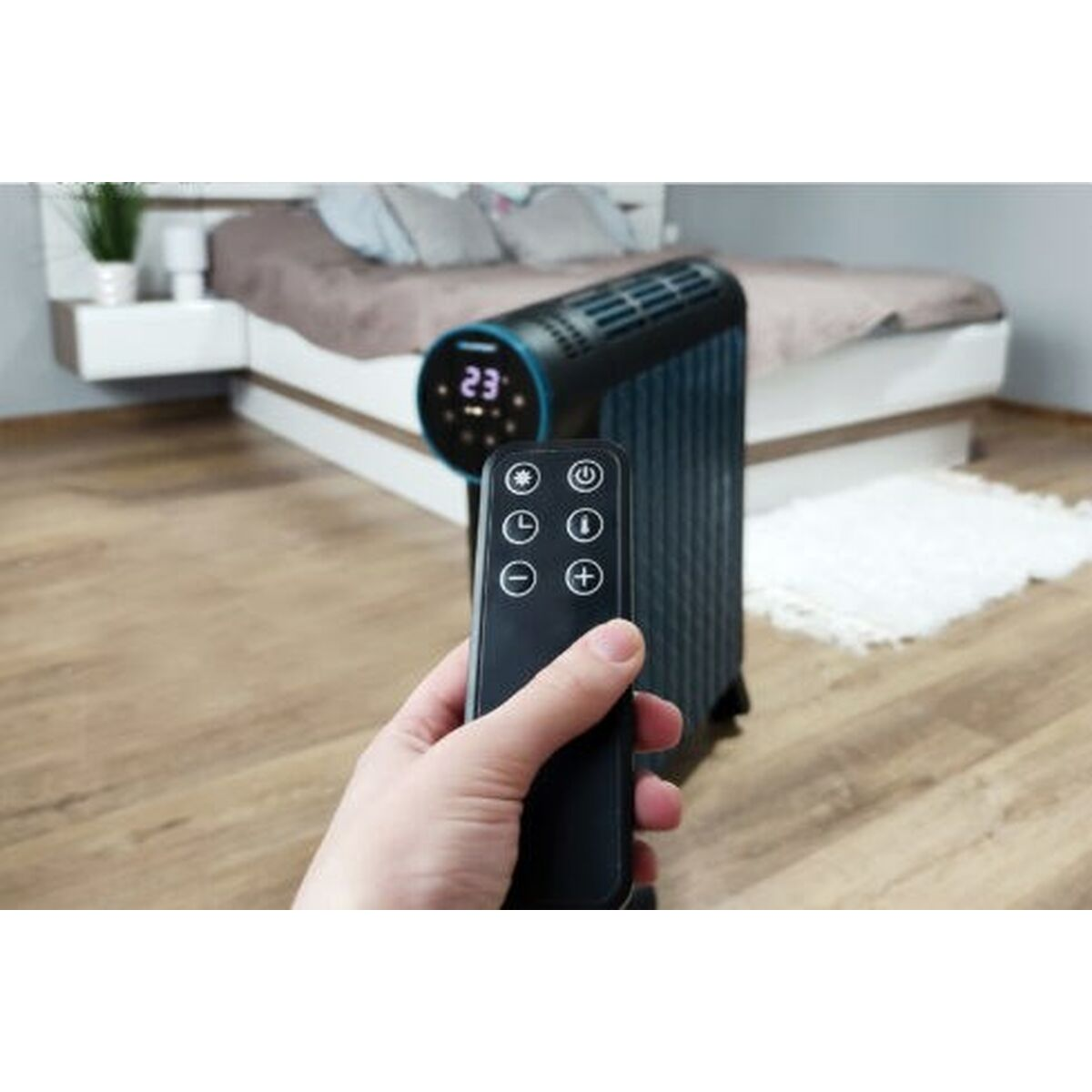 Una mano sosteniendo un mando a distancia negro. Un radiador negro y azul está en el fondo, con una cama.