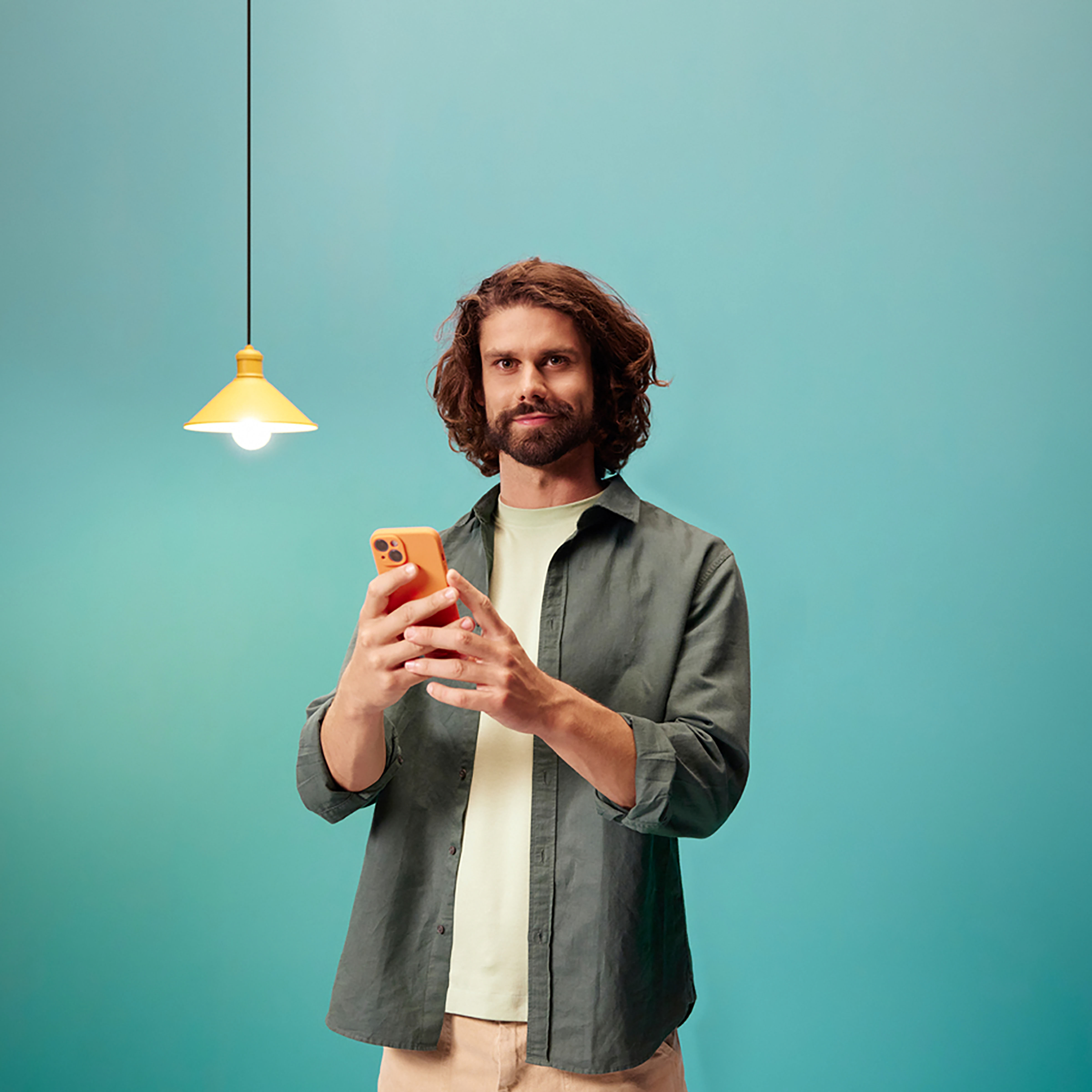 Mężczyzna trzymający pomarańczowy telefon pod żółtą lampą wiszącą na tle turkusowym.