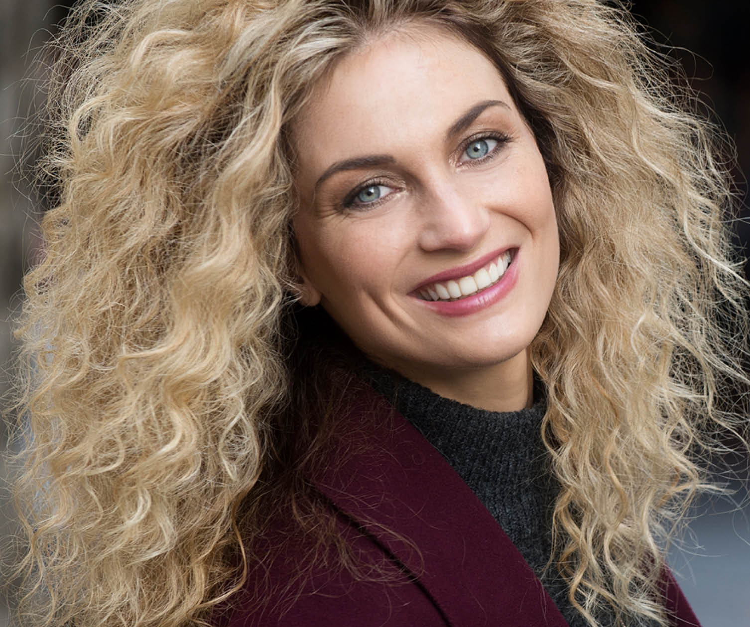 Woman with curly blonde hair smiling, wearing a gray sweater and burgundy coat.