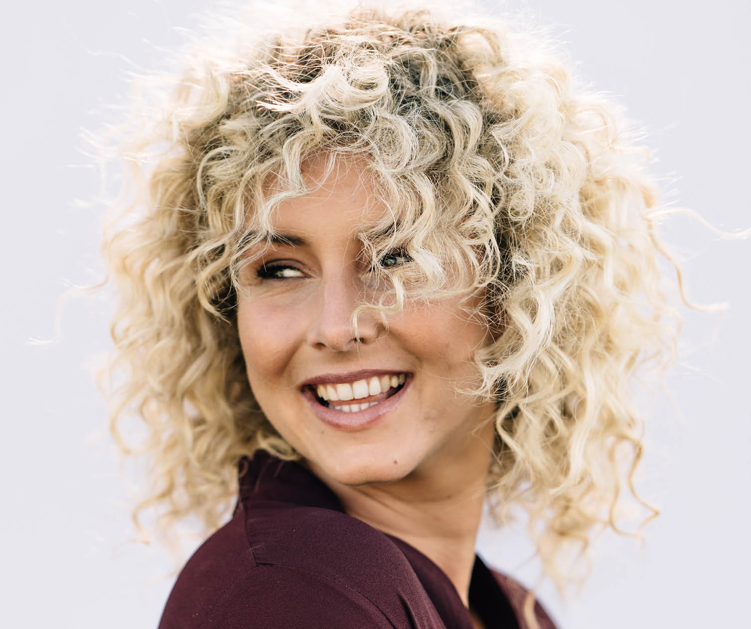 Woman with blonde curly hair smiles, looking back. Wears a dark red top against a white background.