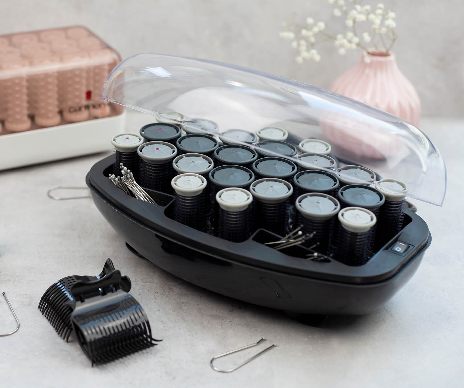 A black hair curler set with rollers, pins and a clear lid. A pink vase is in the background.