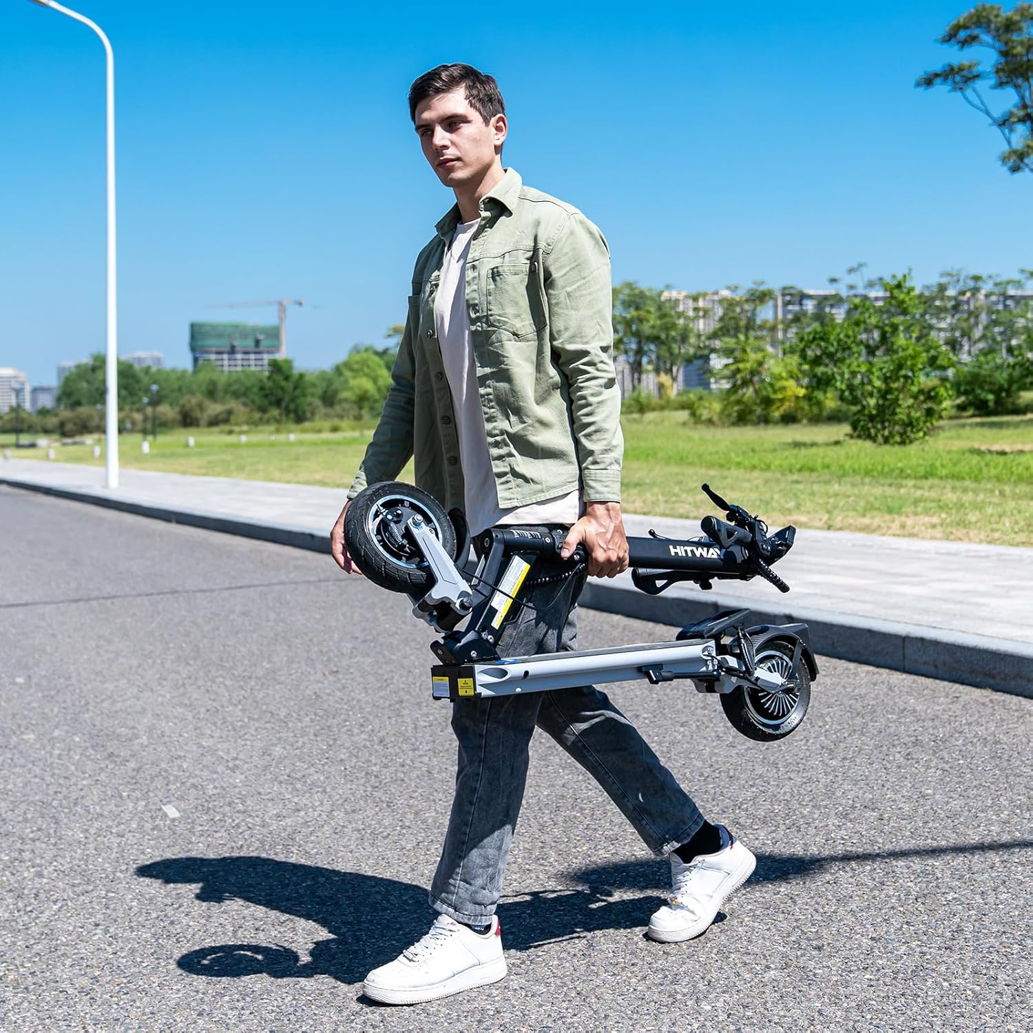 Man carrying a folded e-scooter on a paved road. Sunny day, green trees in the background.