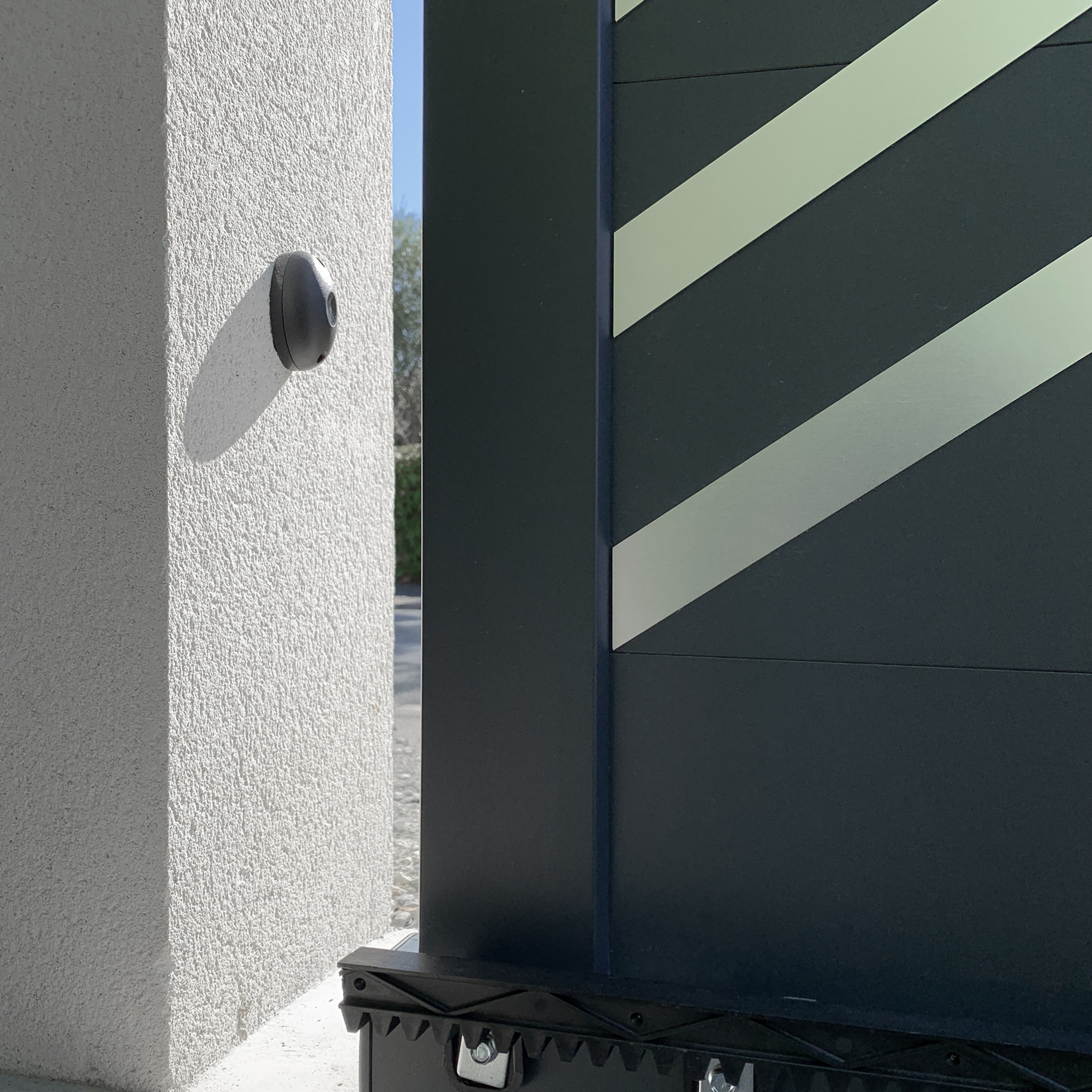 Sensore nero su muro bianco. Cancello nero e argento parzialmente aperto.