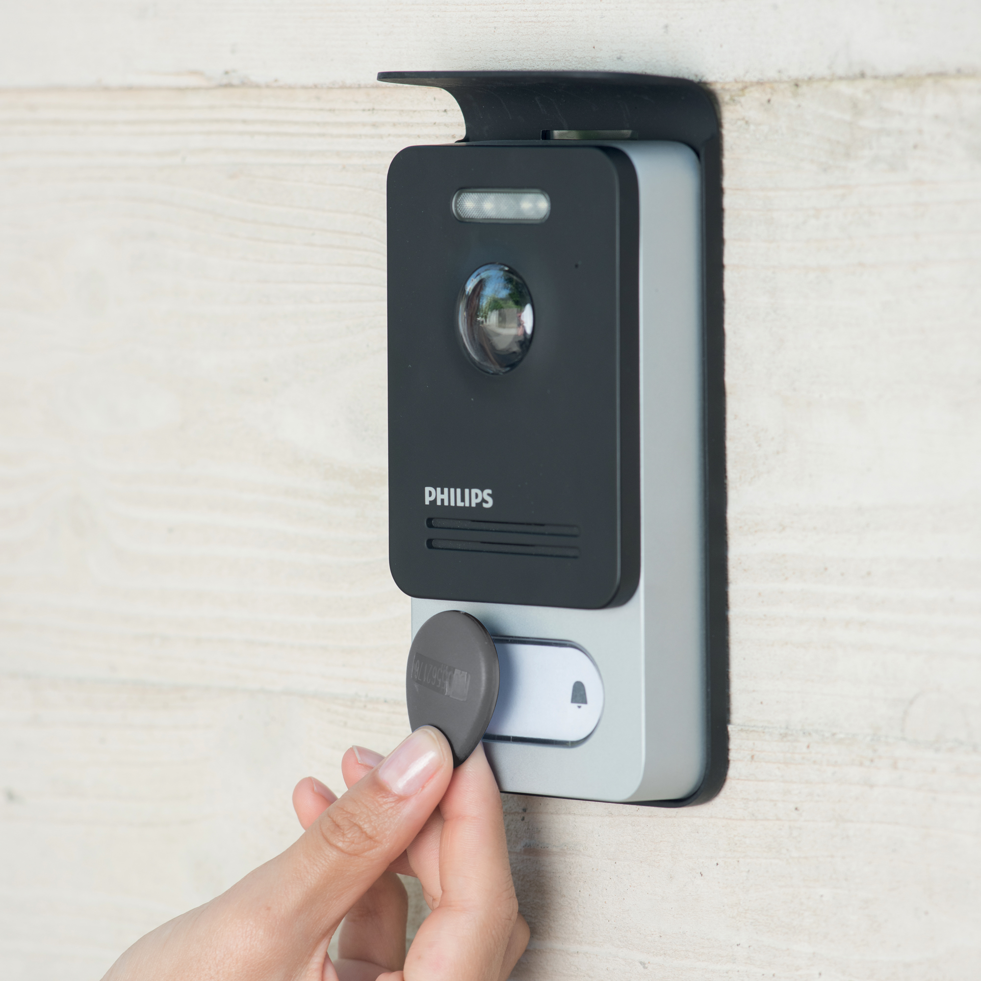 Una mano sosteniendo una tarjeta llave gris cerca de un intercomunicador Philips negro y gris en una pared de madera.