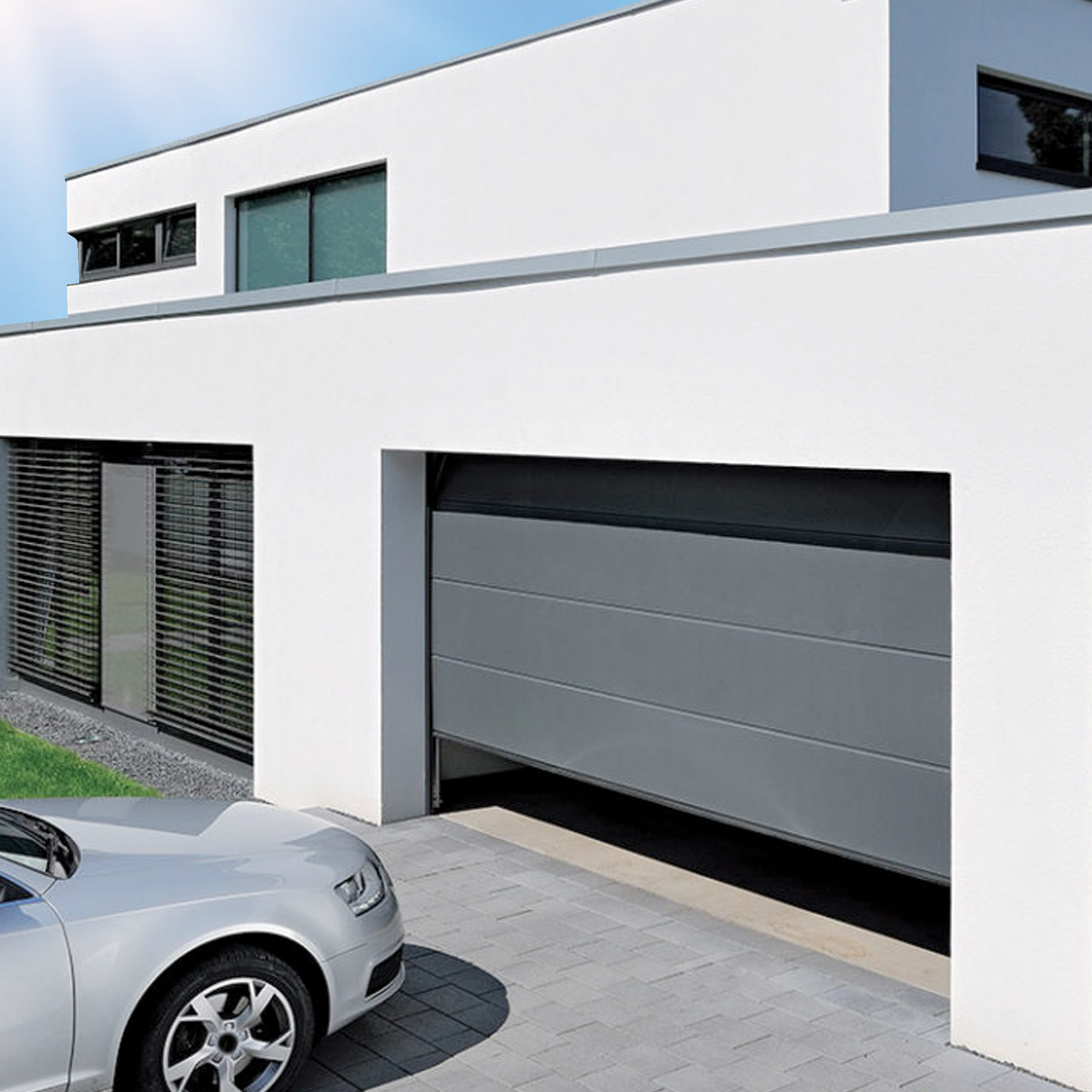 Porta del garage grigia aperta. Edificio bianco con auto parzialmente visibile. Giorno soleggiato.