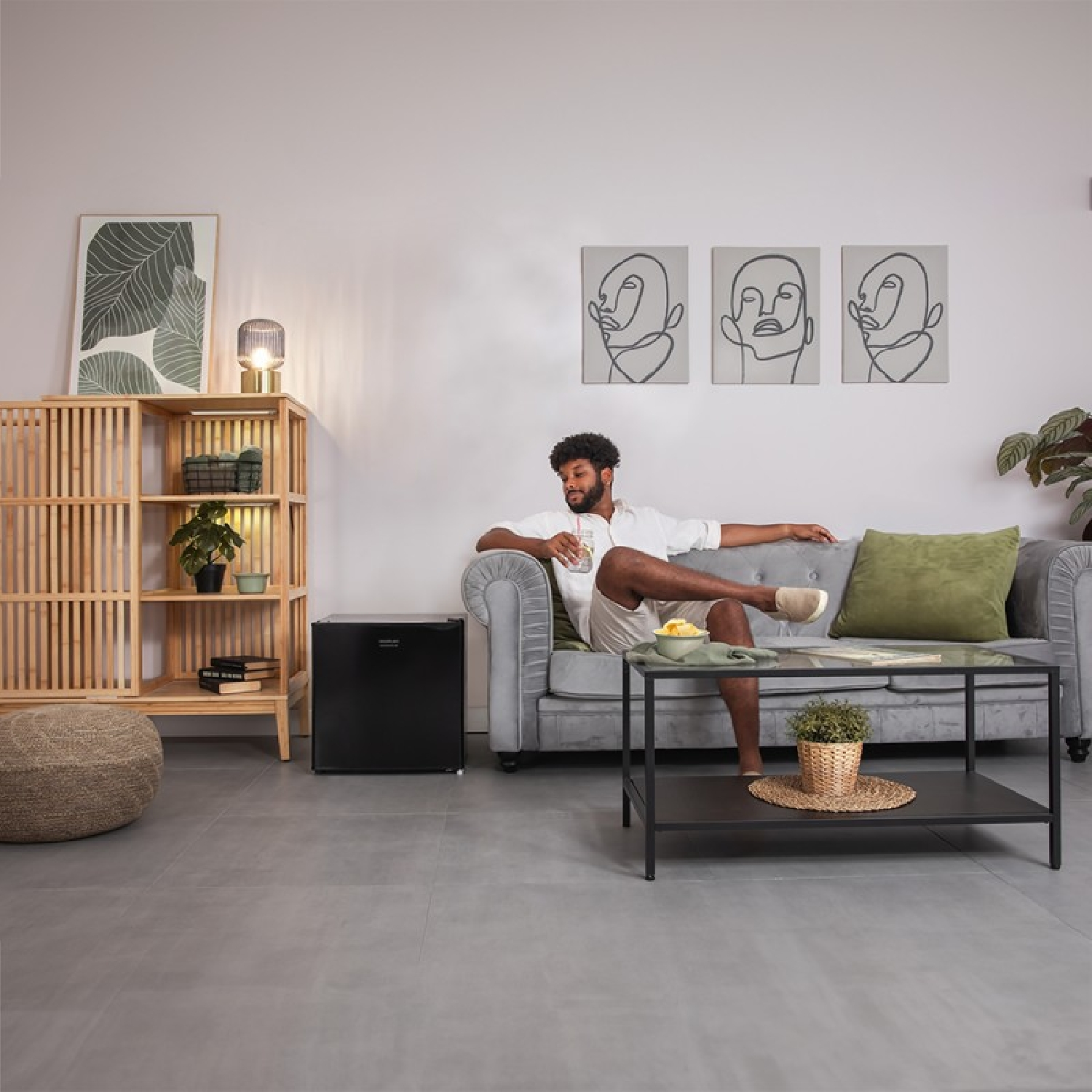 Ein Mann sitzt auf einem grauen Sofa, in der Nähe eines schwarzen Kühlschranks und eines Couchtisches mit Kunst an der Wand.