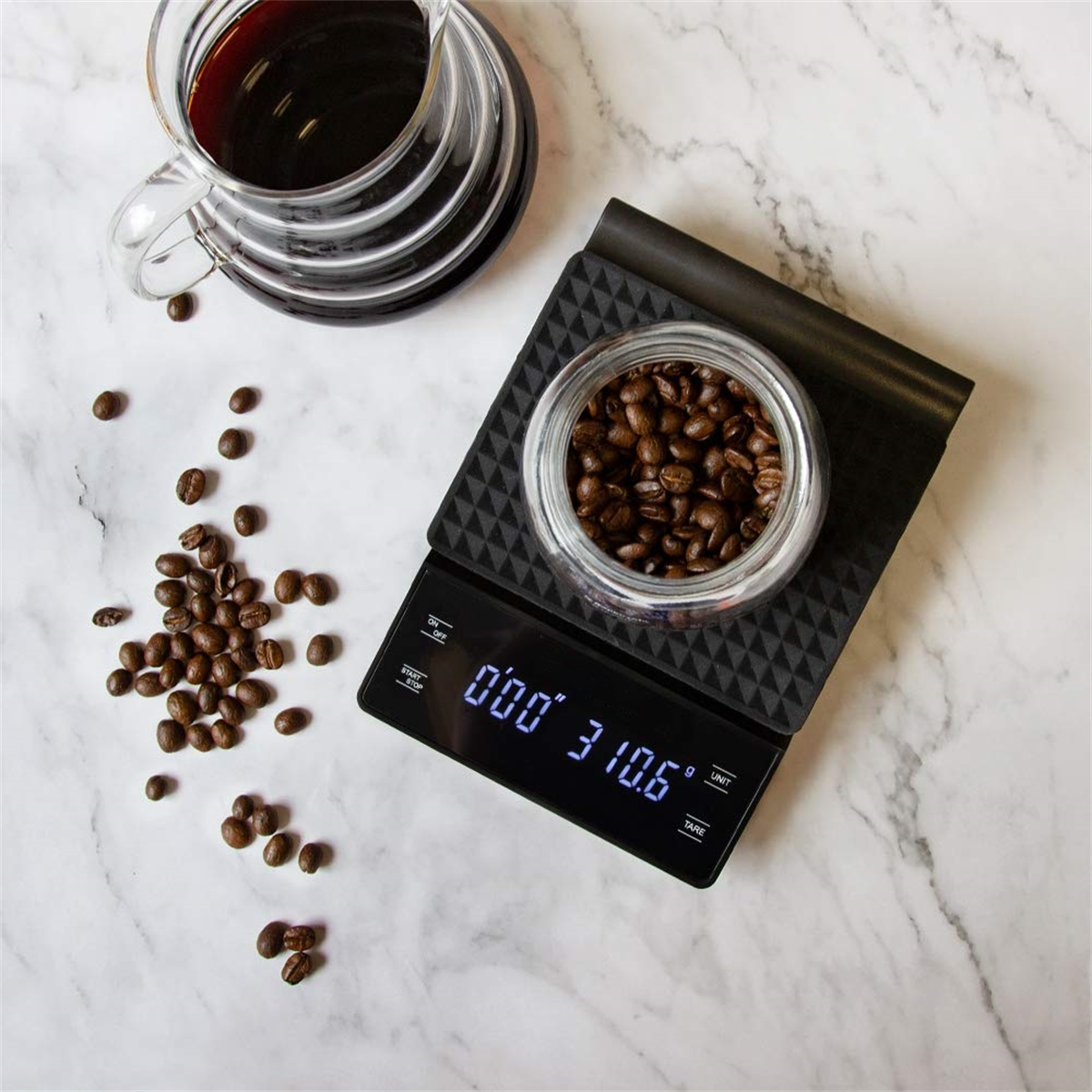 Kaffeebohnen auf einer schwarzen Digitalwaage mit einem Glasbehälter und einer Tasse Kaffee auf Marmoroberfläche.
