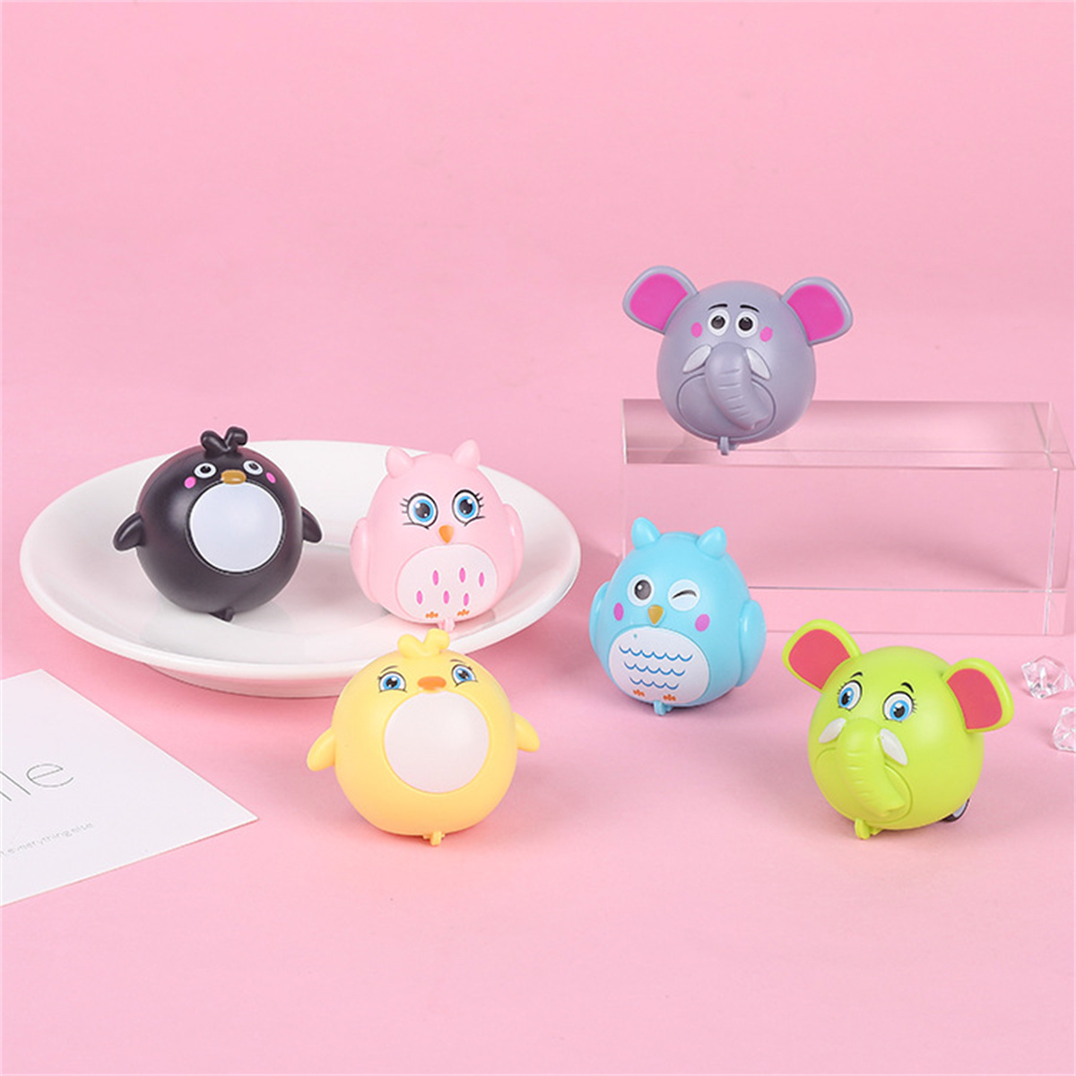 Sechs Spielzeugtiere zum Aufziehen: Pinguin, Eule, Ente und zwei Elefanten, angeordnet auf einer rosa Oberfläche.