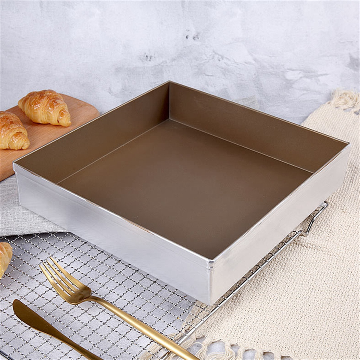 Eine quadratische Backform mit braunem Innenraum auf einem Drahtgestell. Croissants und Besteck liegen daneben.