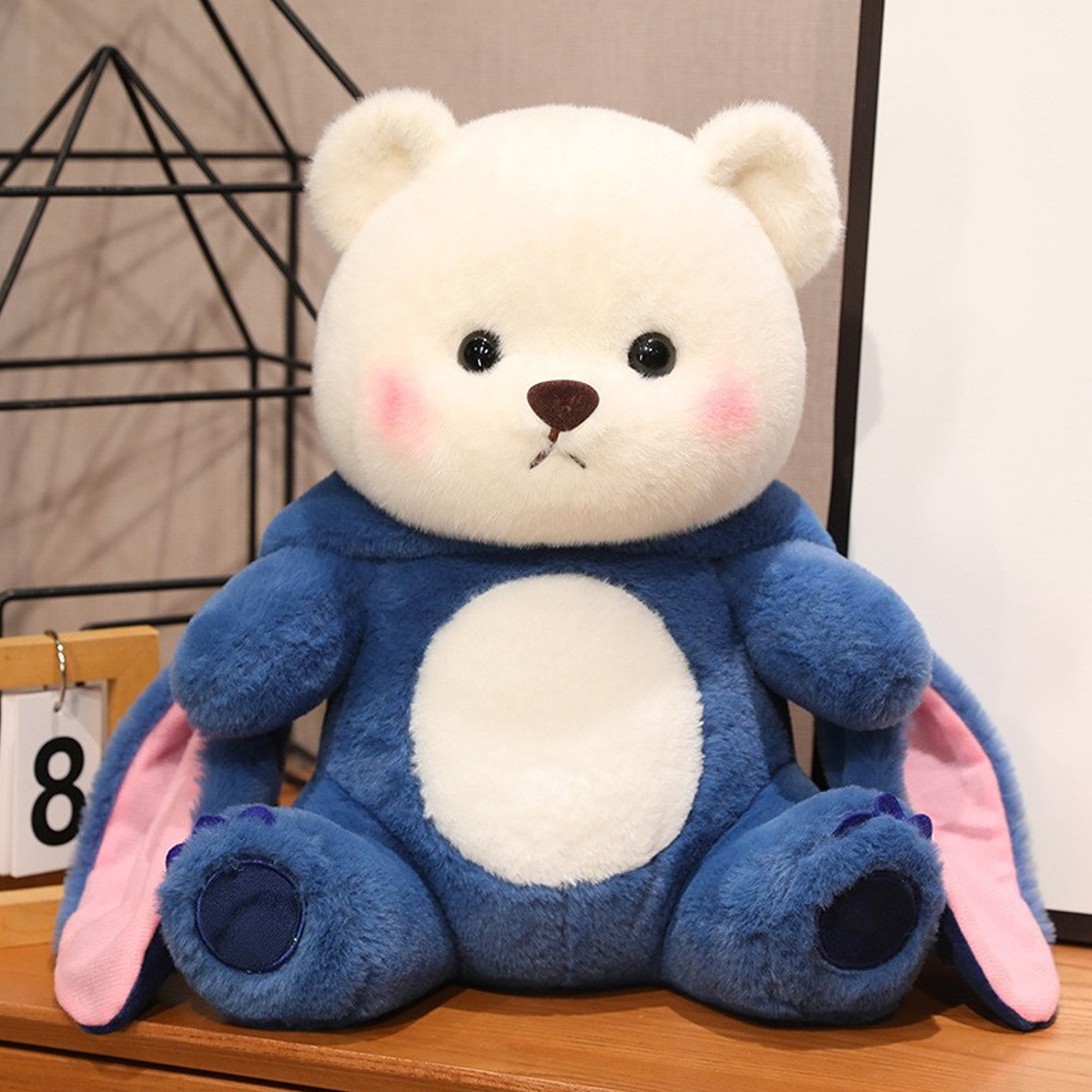 Ein Plüschbär mit weißem Kopf, blauem Körper, rosa Ohren und rosigen Wangen, der auf einer Oberfläche sitzt.