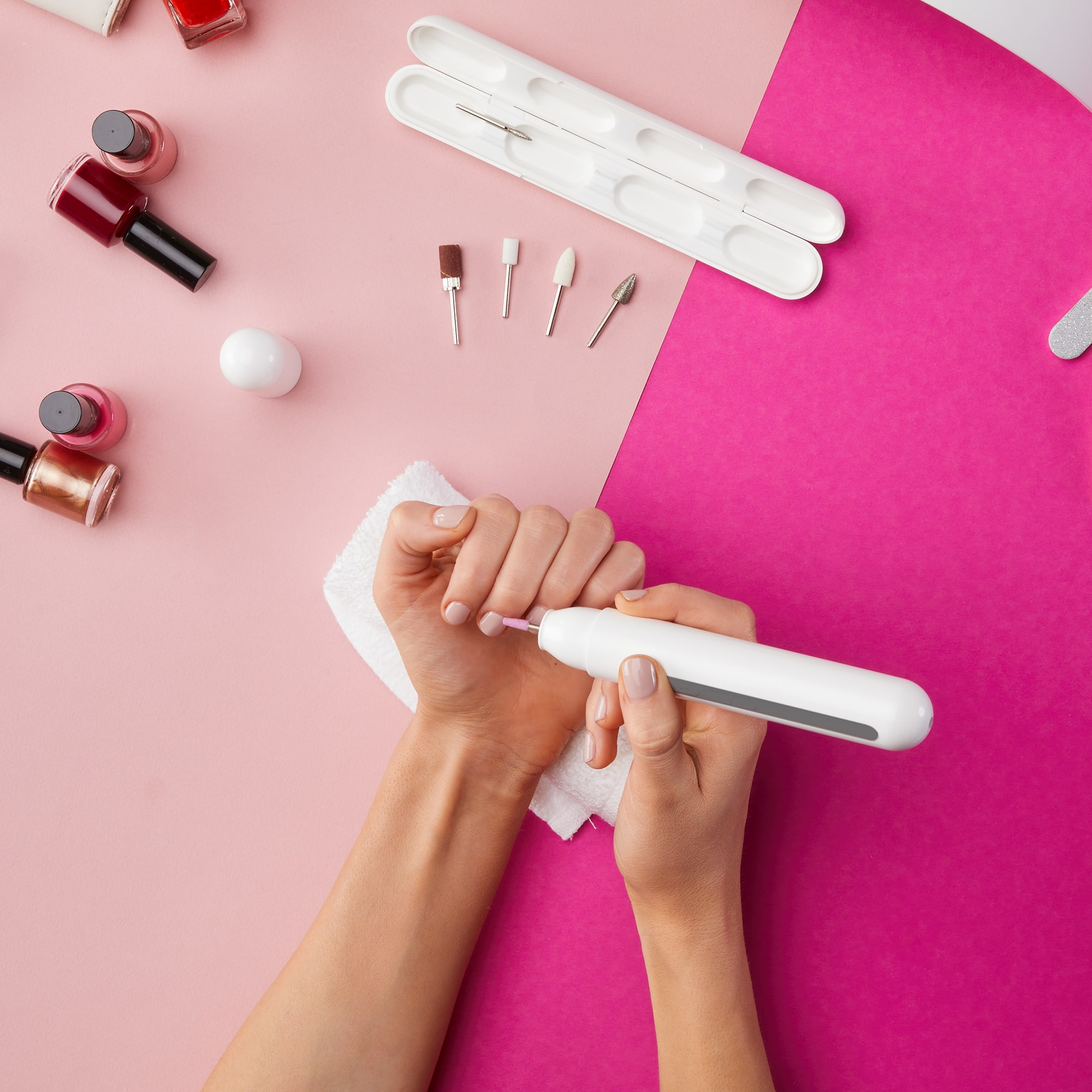 Eine Person benutzt einen Nagelbohrer auf ihren Fingernägeln, mit Nagellackflaschen und Werkzeugen auf rosa Oberfläche.