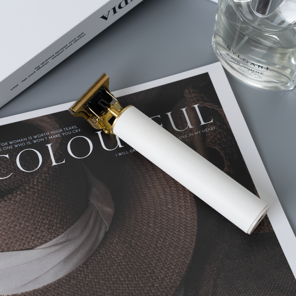 Una recortadora de cabello blanca y dorada sobre una revista con texto y un frasco de perfume al lado.