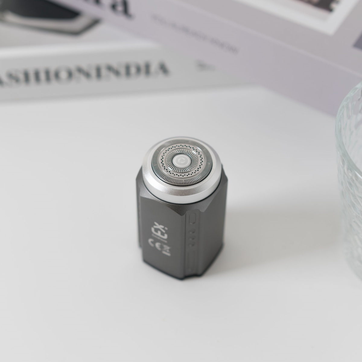 Una afeitadora eléctrica gris y plateada sobre superficie blanca, con un libro y un vaso al fondo.