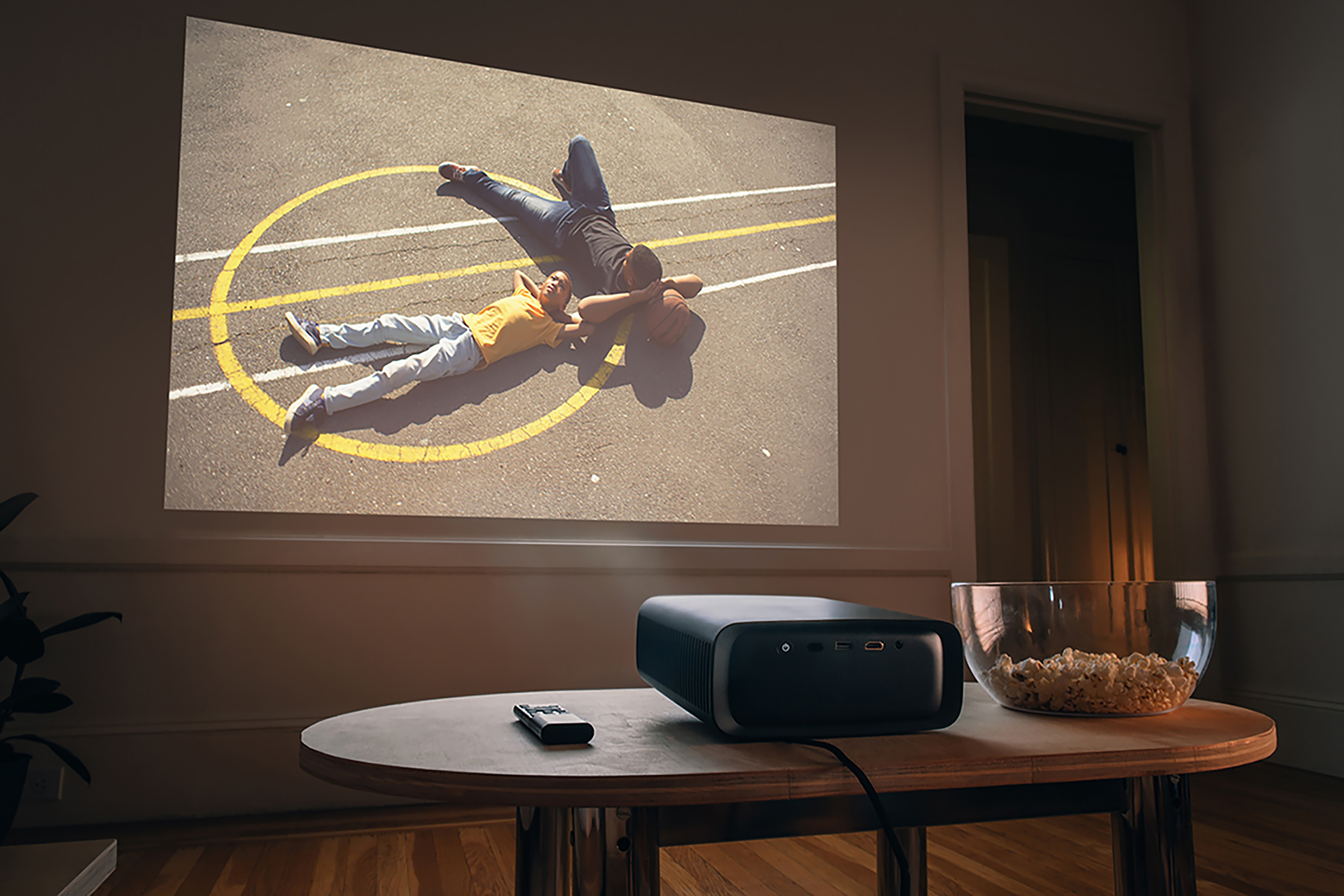 Projektor wyświetla obraz ludzi na ziemi. Miska popcornu na stole, ciemny pokój.