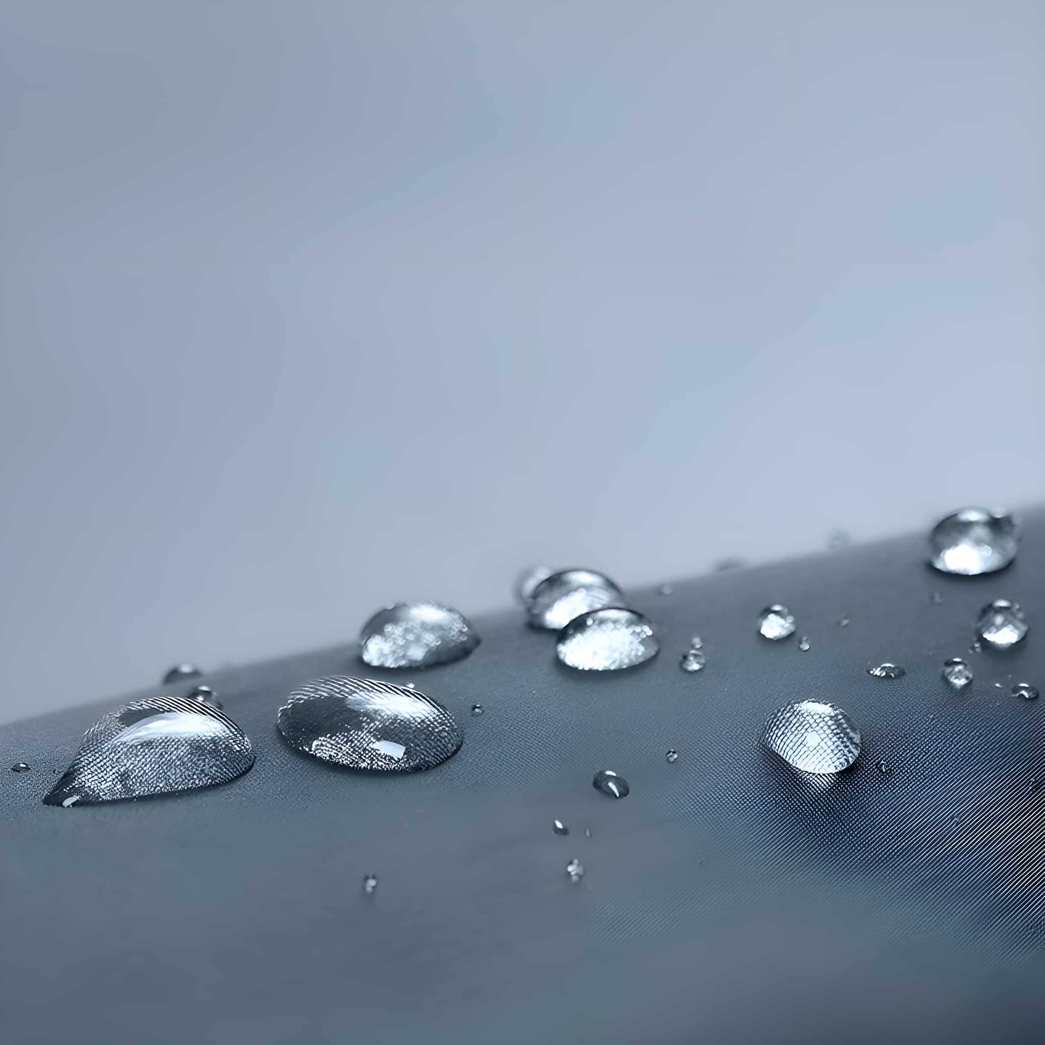 Nahaufnahme von Wassertropfen auf einer grauen, strukturierten Oberfläche. Das Wasser ist klar.