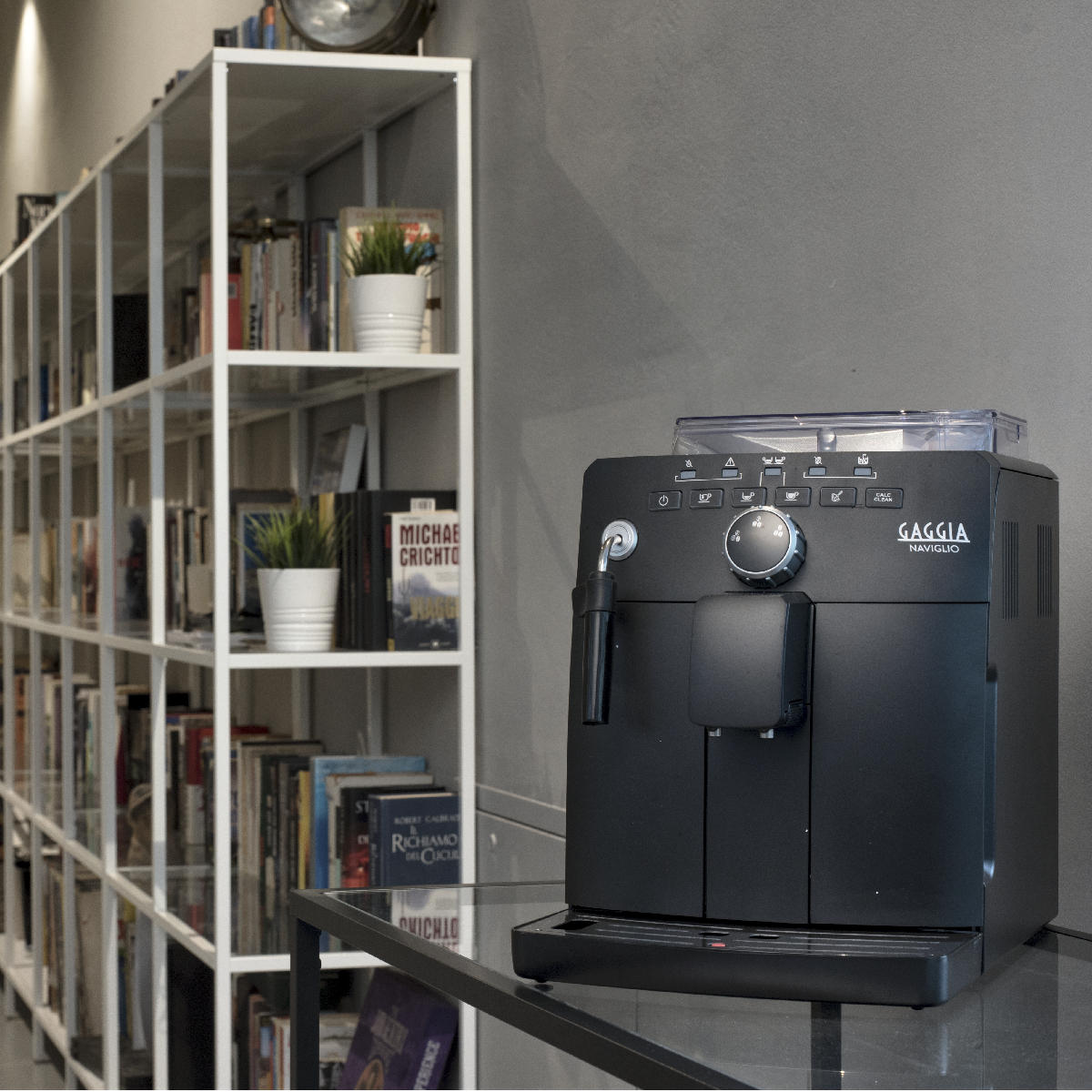 Czarny ekspres do kawy Gaggia Naviglio obok półki na książki i szklanego stołu.